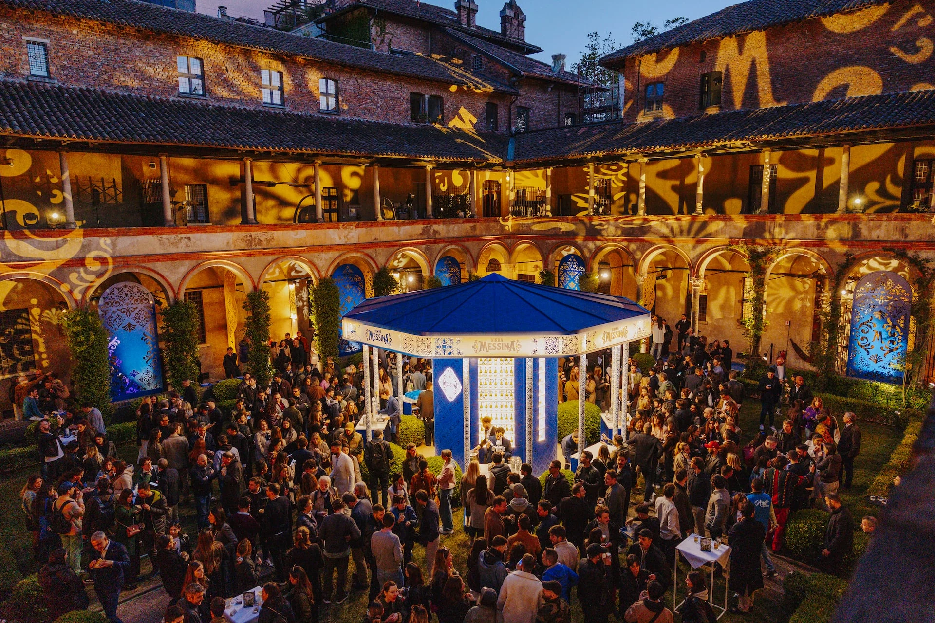 Night view from above of the crowded cloister, arcades and upper walls lit with golden pattern projections – Birra Messina Bar Meraviglia at Fuorisalone, Milan, Italy, 2024 – event design