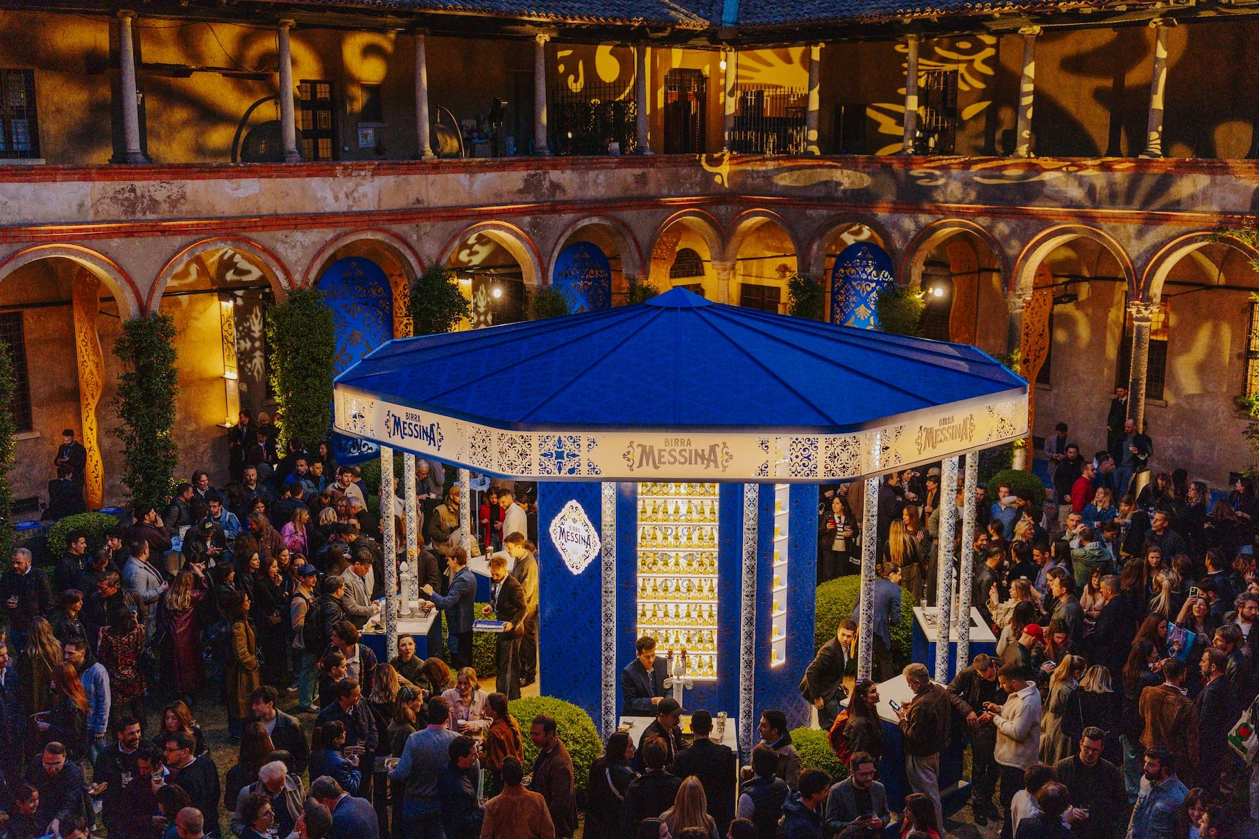 Blue Birra Messina bar kiosk in the crowded cloister at dusk, arcades washed with golden pattern projections – Birra Messina Bar Meraviglia at Fuorisalone, Milan, Italy, 2024 – event design