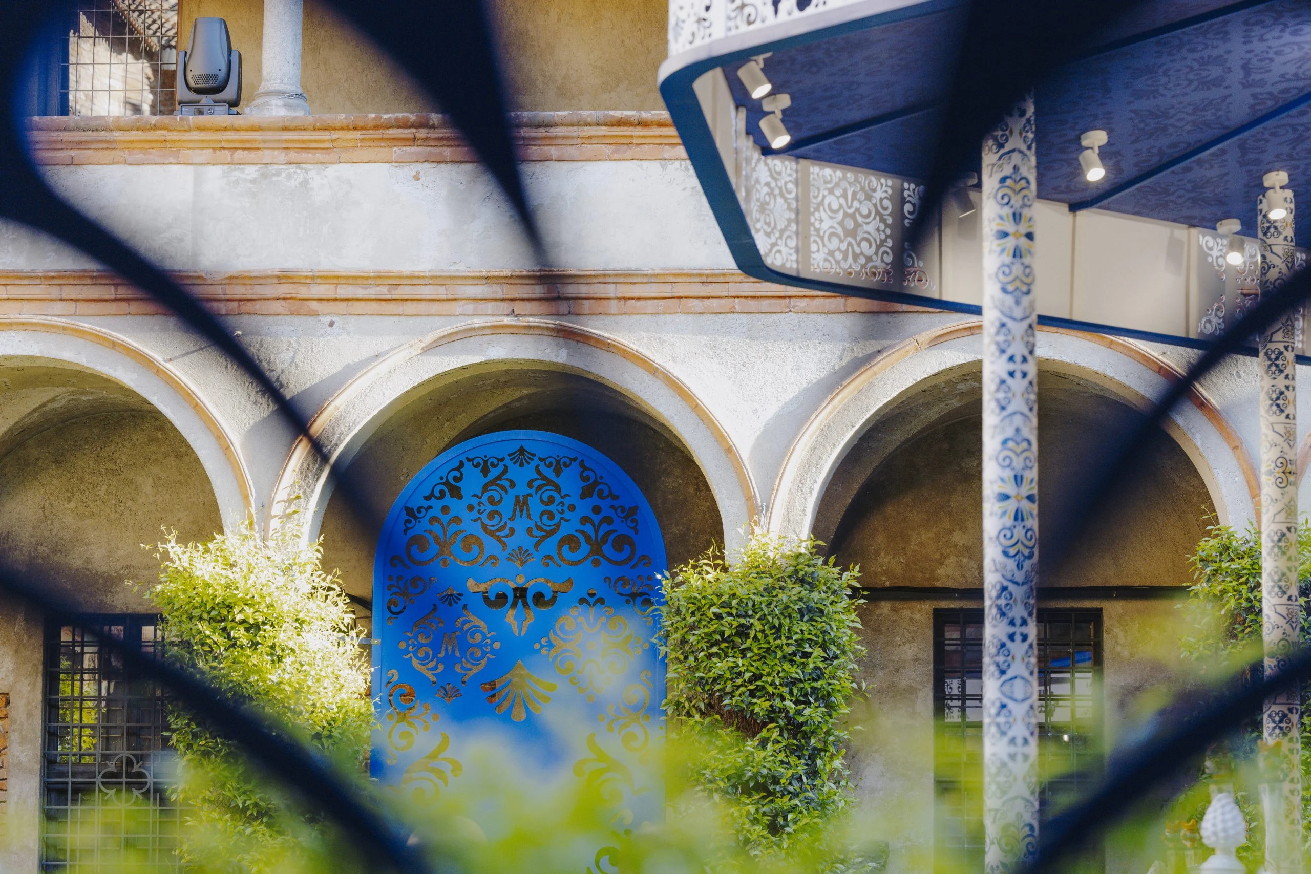 Blue ornamental cut-out panel set in a cloister arch, seen through blurred blue shapes in the foreground – Birra Messina Bar Meraviglia at Fuorisalone, Milan, Italy, 2024 – event design