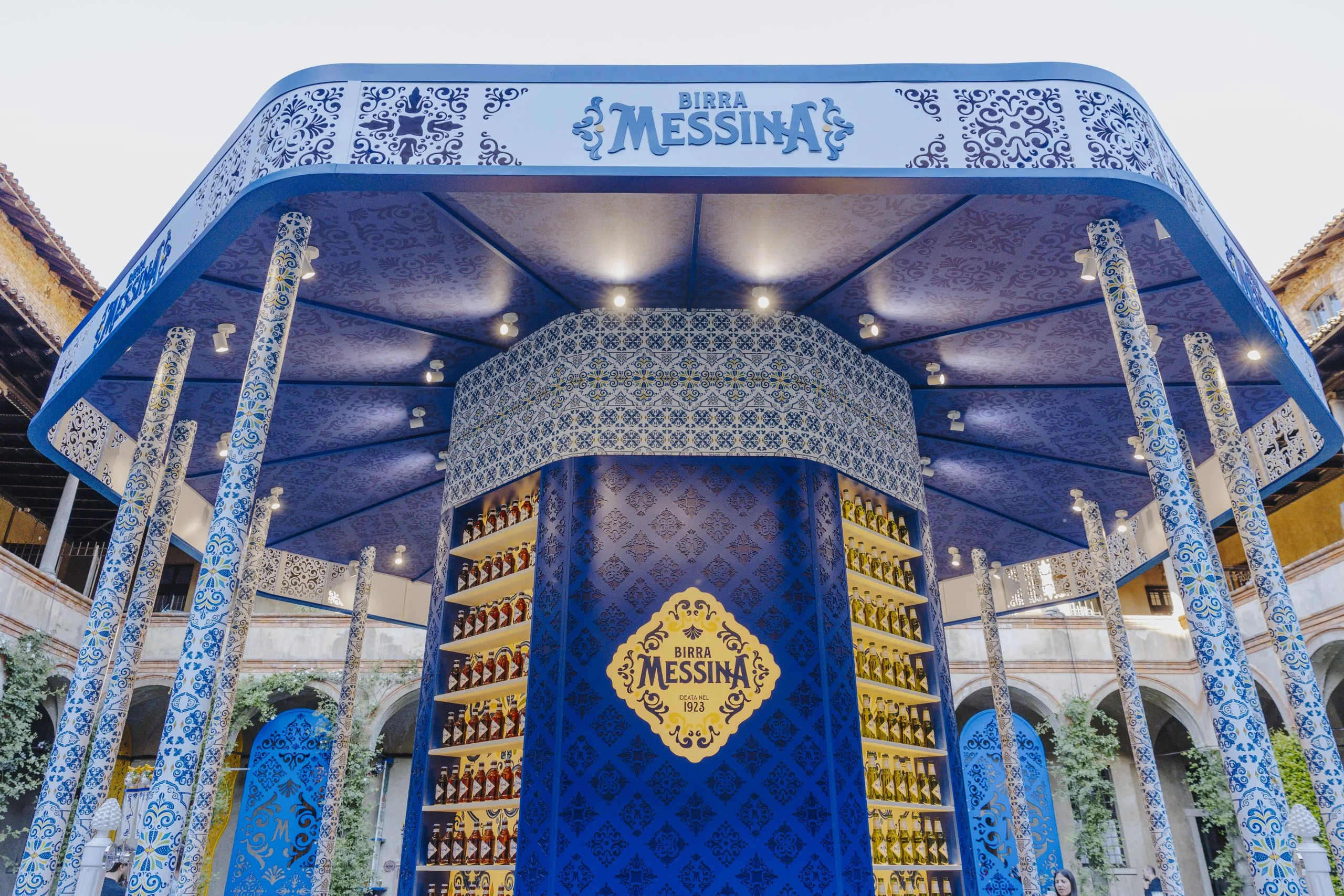 Close view of the Birra Messina kiosk: tiled columns, lit canopy and a central tower of beer bottles – Birra Messina Bar Meraviglia at Fuorisalone, Milan, Italy, 2024 – event design