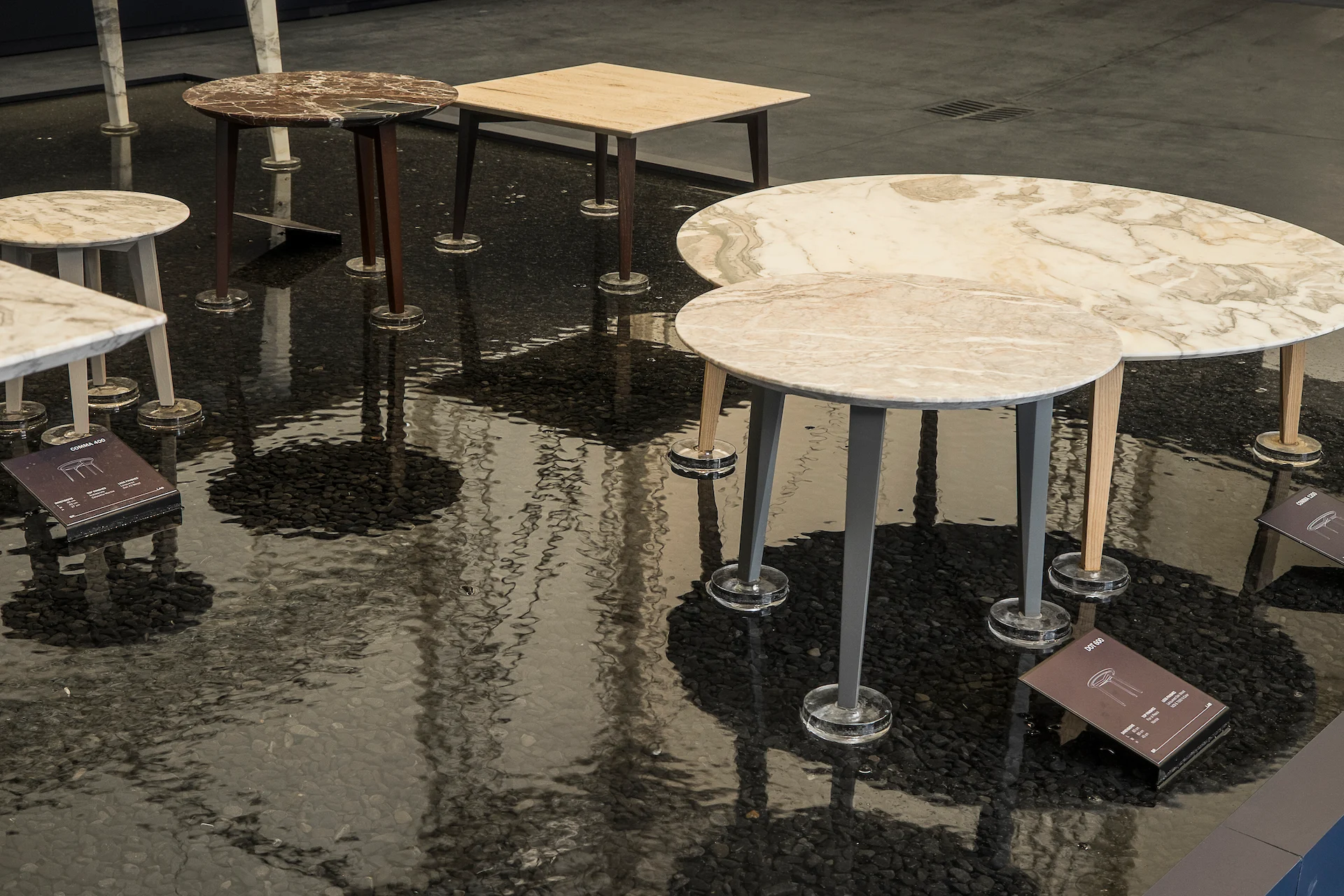 Round marble and wood side tables with small info plaques standing in shallow dark water – Factory Reflections at Sice Previt, Segrate, Italy, 2024 – exhibition design
