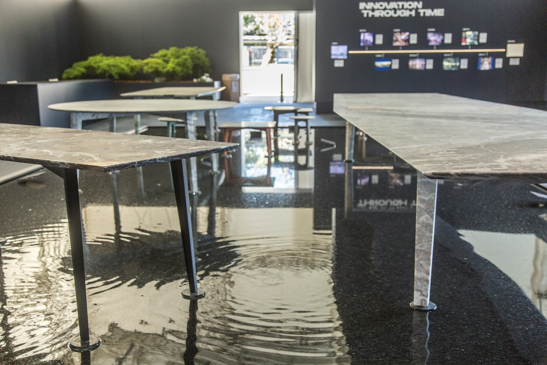 Marble tables rising from rippling water, with a moss planter and the Innovation Through Time wall behind – Factory Reflections at Sice Previt, Segrate, Italy, 2024 – exhibition design