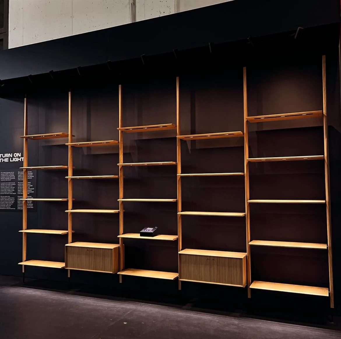 Wall-mounted wooden shelving system with cabinets lit against a dark wall, beside the Turn on the Light panel – Factory Reflections at Sice Previt, Segrate, Italy, 2024 – exhibition design