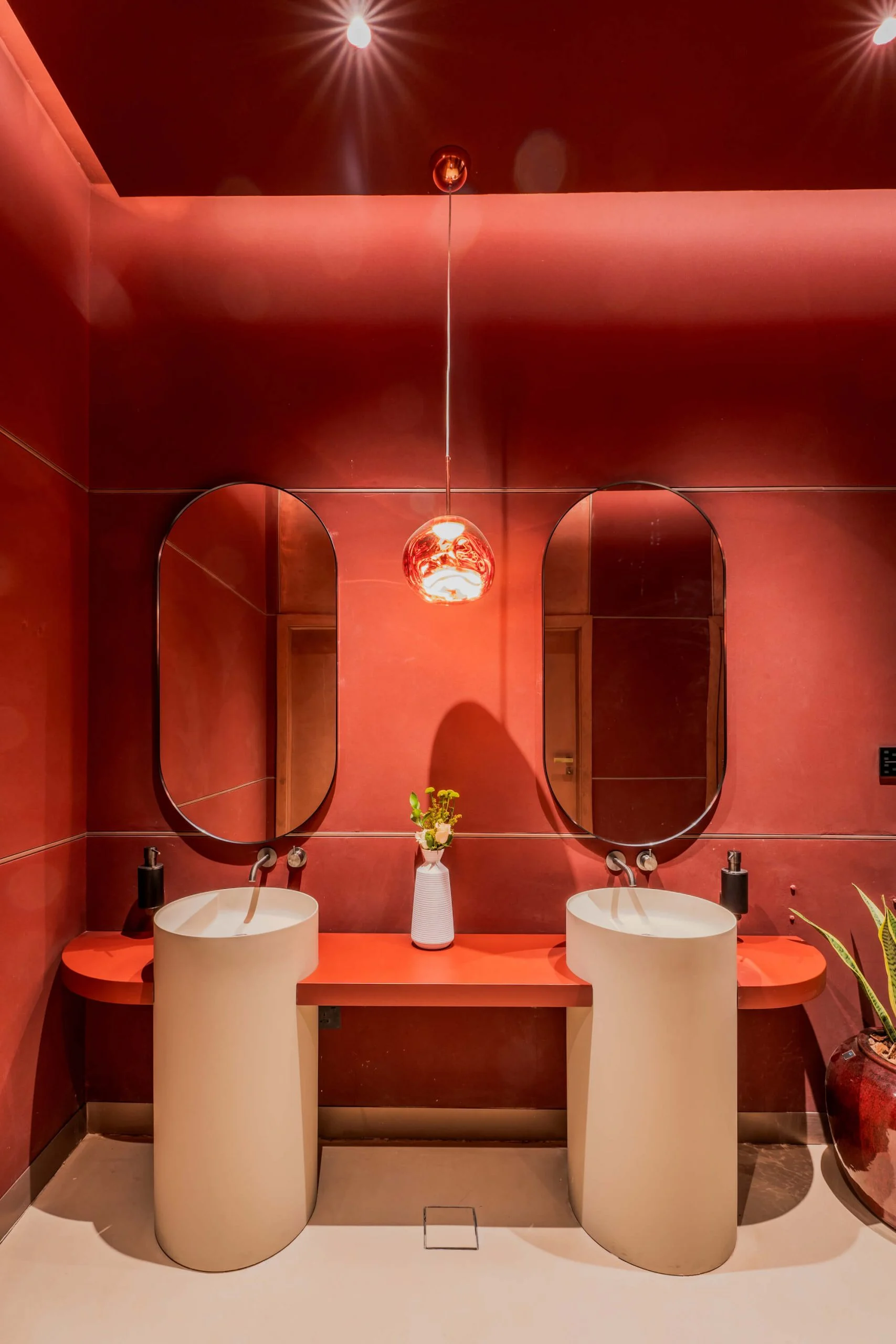 Frontal view of the red restroom: twin white pedestal basins below oval mirrors and a copper pendant lamp – King Fahad Cultural Center, Riyadh, Saudi Arabia, 2024 – architectural renovation