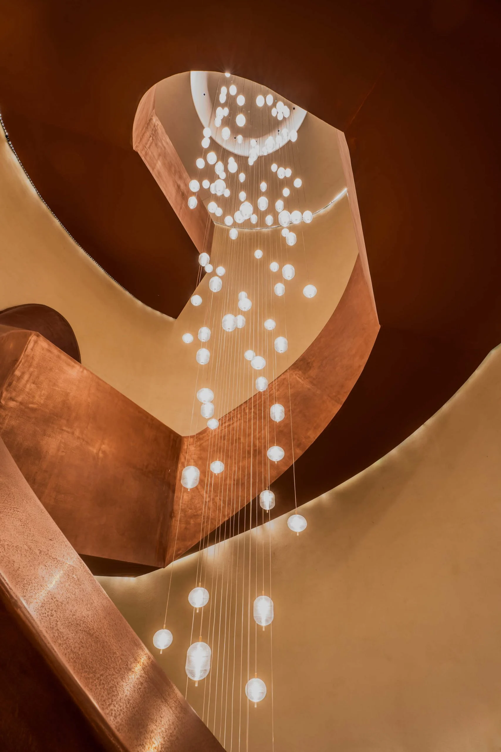 Looking up the copper spiral staircase at a cascade of suspended glass globe lights – King Fahad Cultural Center, Riyadh, Saudi Arabia, 2024 – architectural renovation