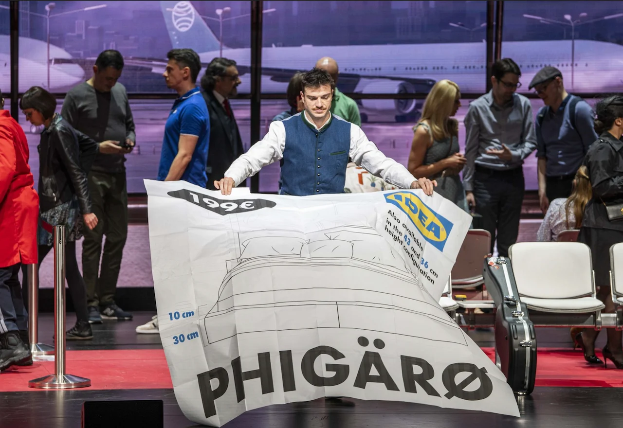 Singer holding up a giant furniture-catalogue sheet labelled Phigärø in the airport terminal – Une Folle Journée Mozart Gala, Salzburg, Austria, 2024 – opera set design
