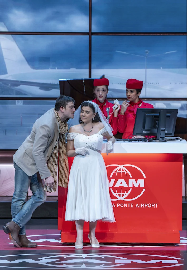Bride in white leaning on the WAM check-in desk while a man whispers, flight attendants behind – Une Folle Journée Mozart Gala, Salzburg, Austria, 2024 – opera set design