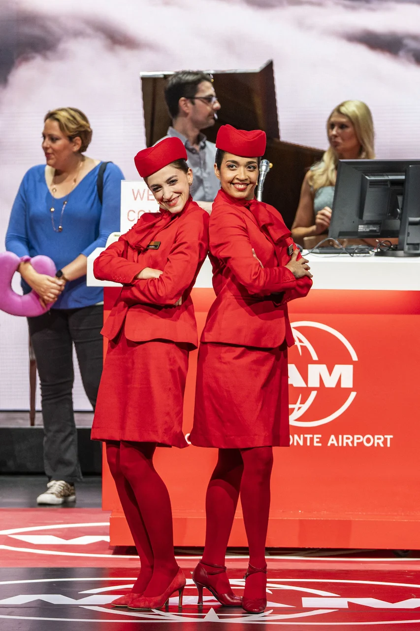 Two performers in red flight attendant uniforms posing back to back by the WAM airport desk – Une Folle Journée Mozart Gala, Salzburg, Austria, 2024 – opera set design