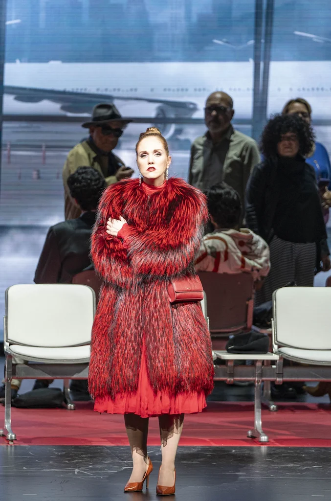 Singer in a red fur coat standing in front of rows of airport seats and passengers – Une Folle Journée Mozart Gala, Salzburg, Austria, 2024 – opera set design