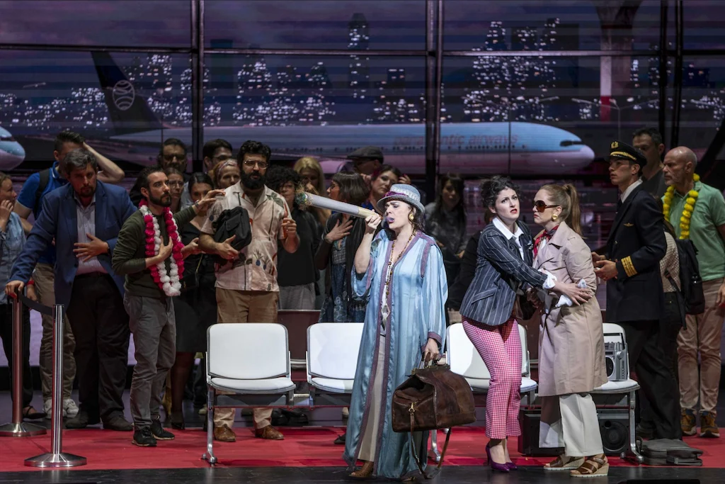 Singer in a blue robe and hat with a travel bag, surrounded by passengers in the terminal at night – Une Folle Journée Mozart Gala, Salzburg, Austria, 2024 – opera set design