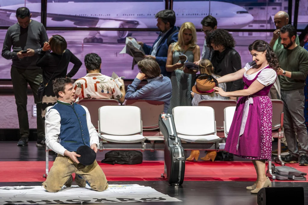 Singer in a waistcoat kneeling before a singer in a pink dirndl among airport seats – Une Folle Journée Mozart Gala, Salzburg, Austria, 2024 – opera set design