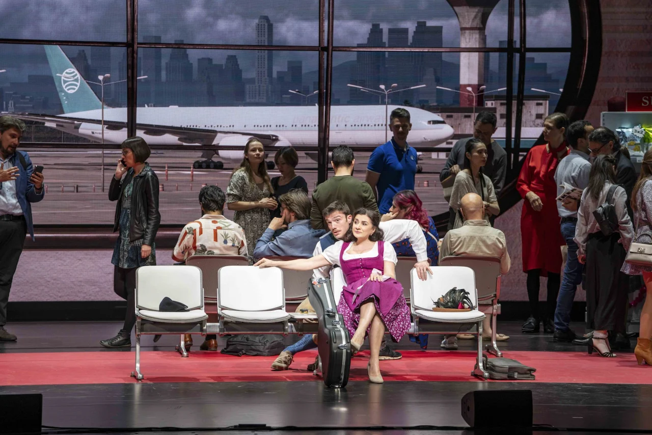 Singer in a pink dress posing on airport seats with a cello case, passengers and a plane behind – Une Folle Journée Mozart Gala, Salzburg, Austria, 2024 – opera set design