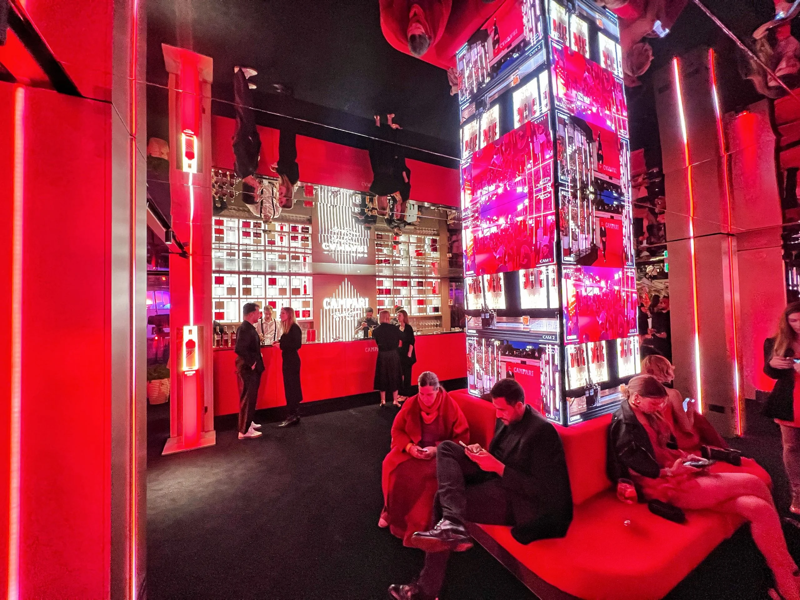 Red-lit lounge at night with guests on red sofas, a mirrored ceiling and a column clad in video screens – Campari Cannes Film Festival 2024, France – event architecture