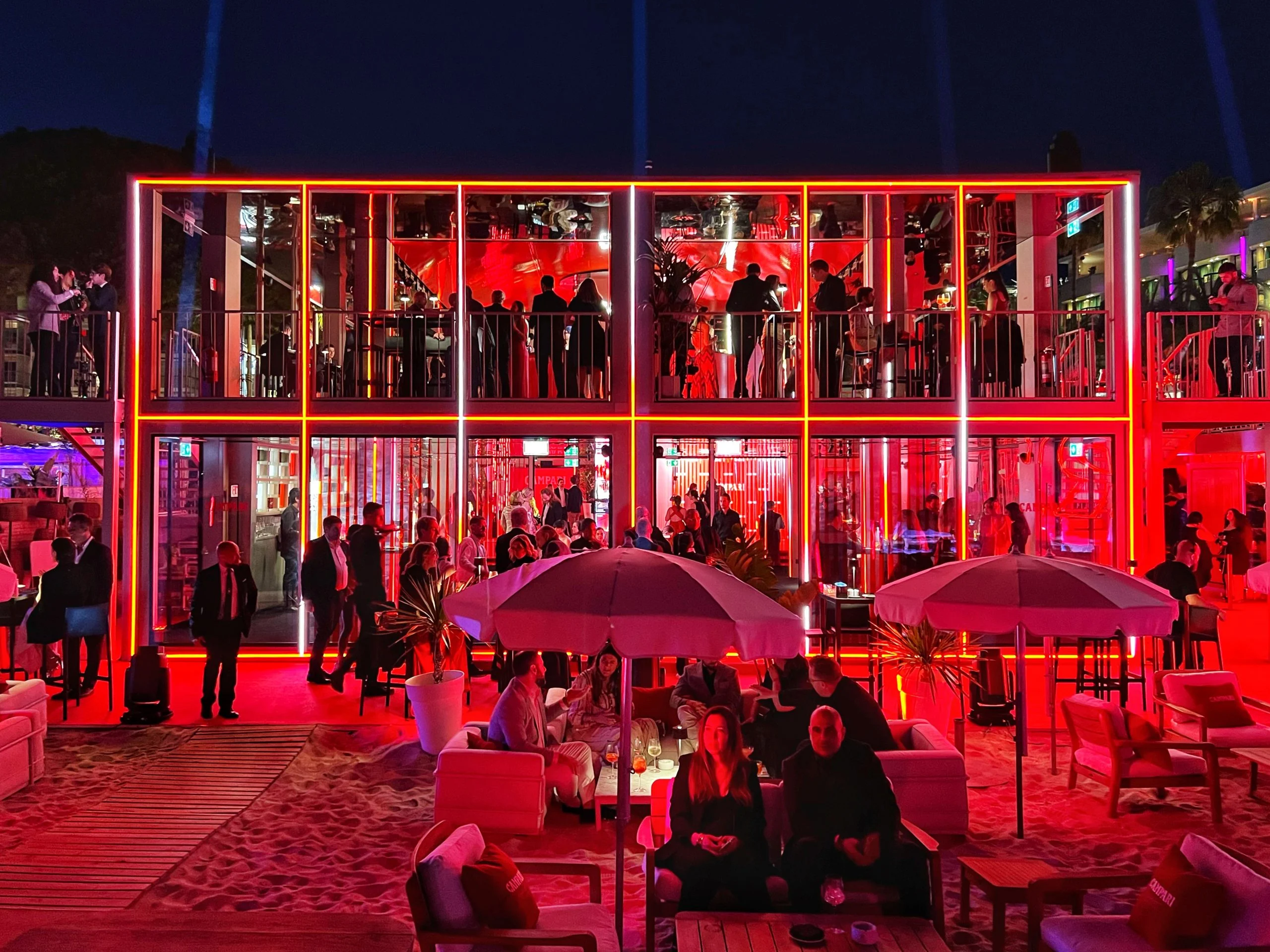 Pavilion facade glowing red at night, guests on both floors and under umbrellas on the sand – Campari Cannes Film Festival 2024, France – event architecture