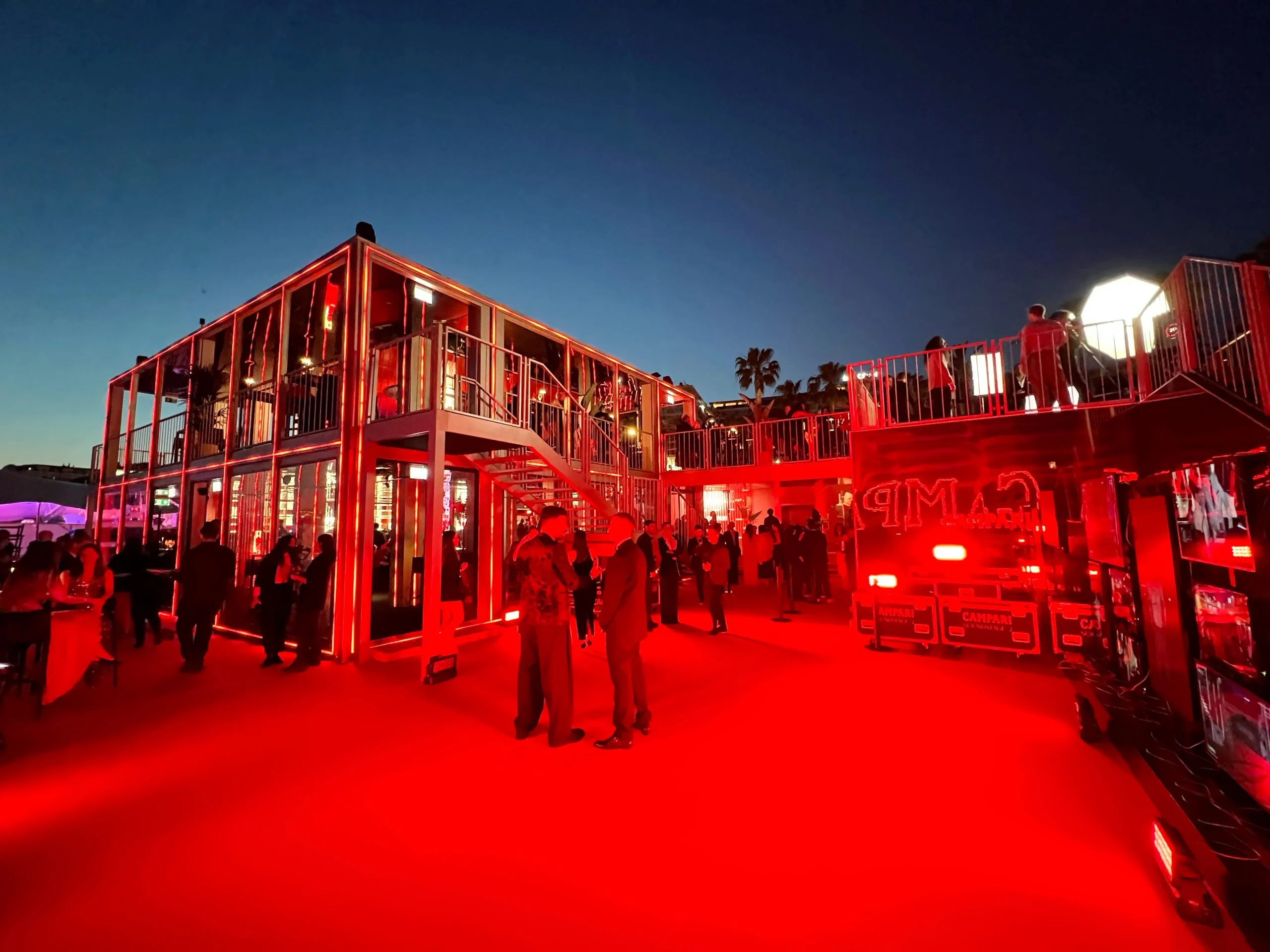 Red-lit forecourt at dusk with the two-storey glass pavilion, its outdoor stairs and a red terrace – Campari Cannes Film Festival 2024, France – event architecture