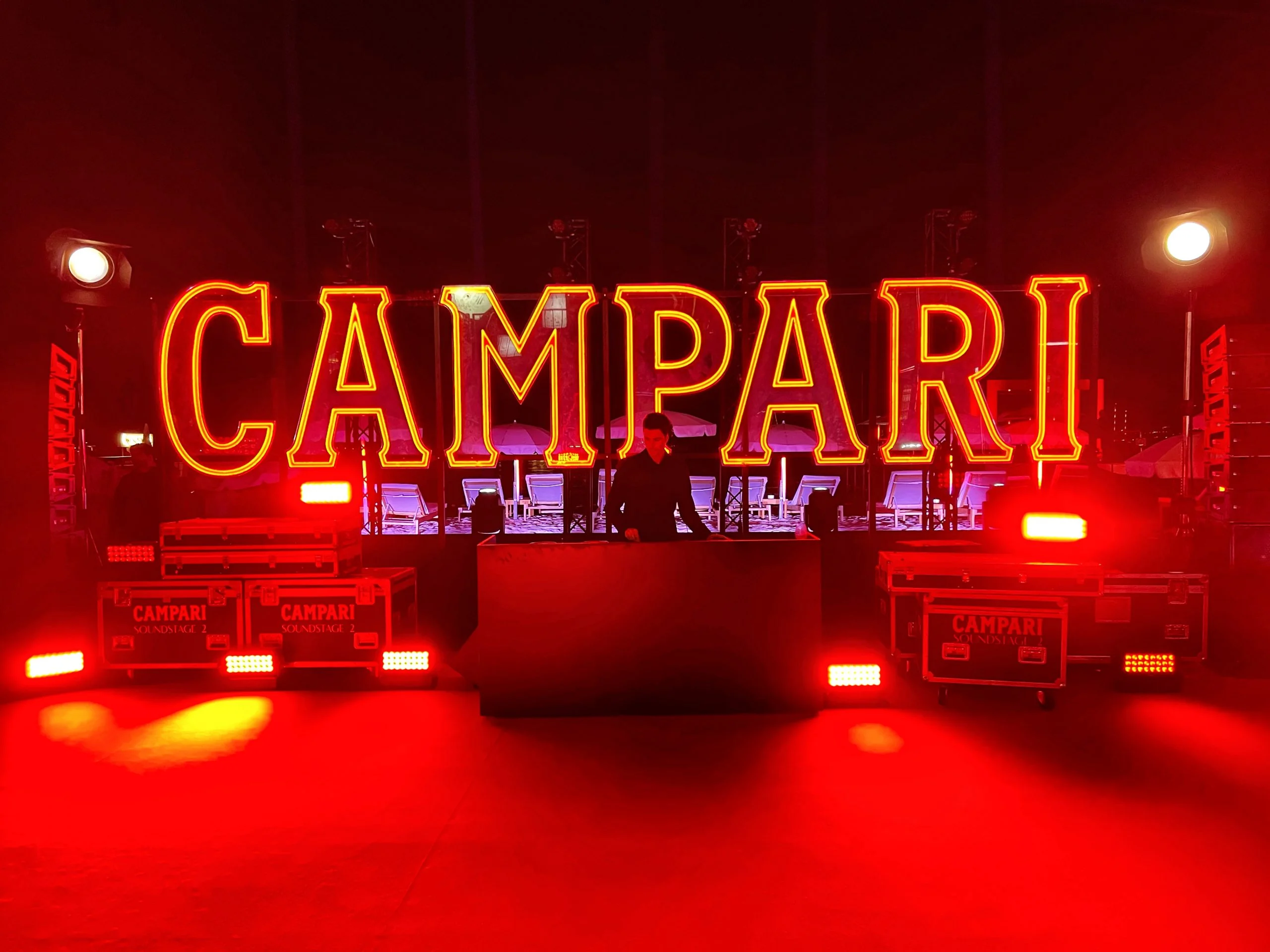 Giant illuminated Campari letters above a DJ booth, flanked by flight cases and red lights – Campari Cannes Film Festival 2024, France – event architecture