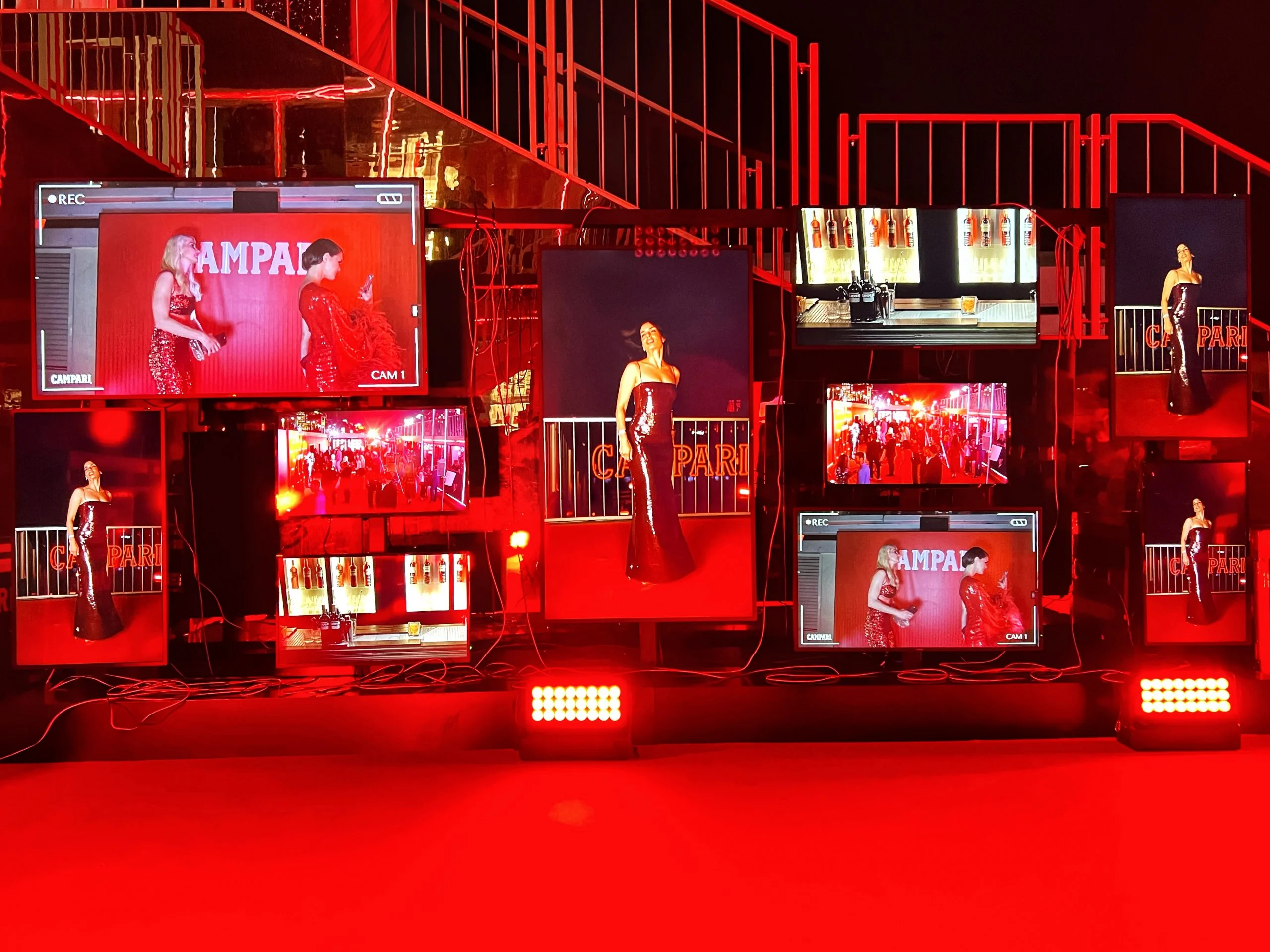 Wall of screens showing live camera feeds of guests posing against a Campari backdrop, red-lit stairs behind – Campari Cannes Film Festival 2024, France – event architecture
