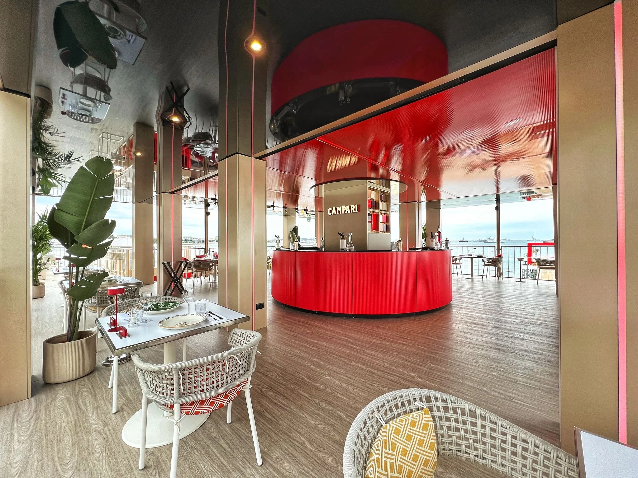 Terrace dining area with wicker chairs and plants, looking towards the round red Campari bar – Campari Cannes Film Festival 2024, France – event architecture