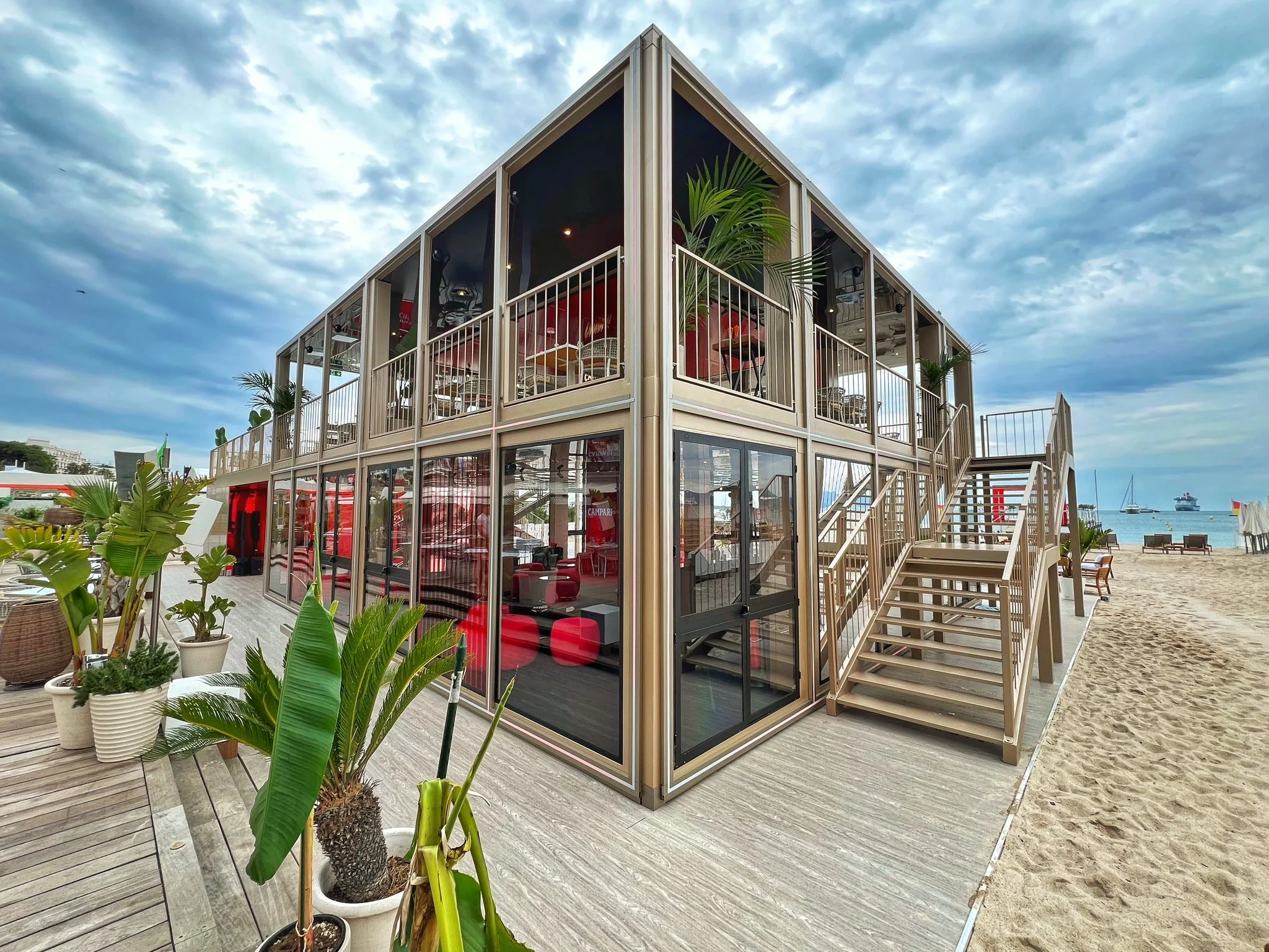 Two-storey glass and champagne-gold pavilion on the Cannes beach, with timber stairs and potted palms – Campari Cannes Film Festival 2024, France – event architecture
