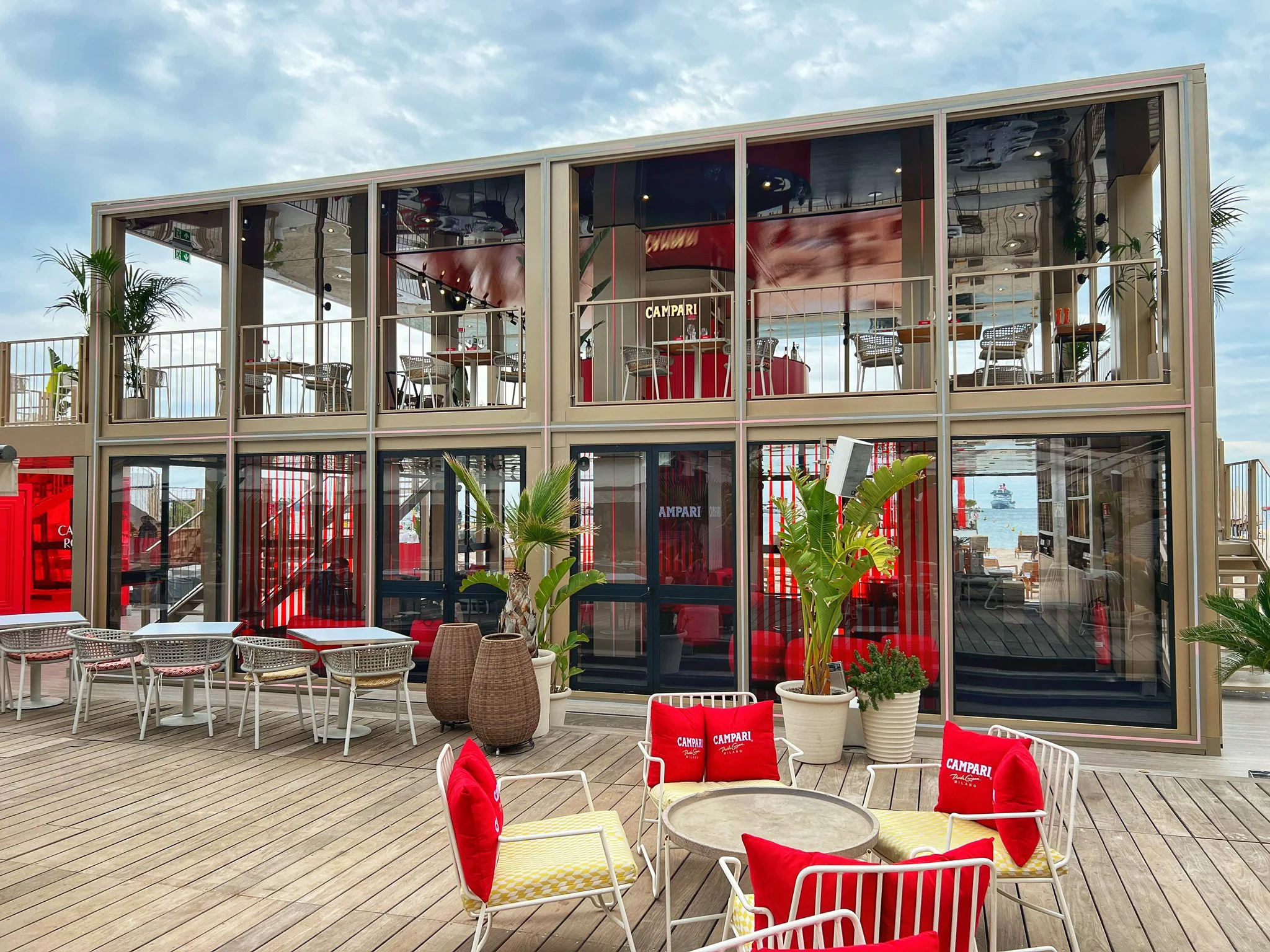 Glazed facade of the Campari pavilion with red interiors, deck seating with red Campari cushions in front – Campari Cannes Film Festival 2024, France – event architecture