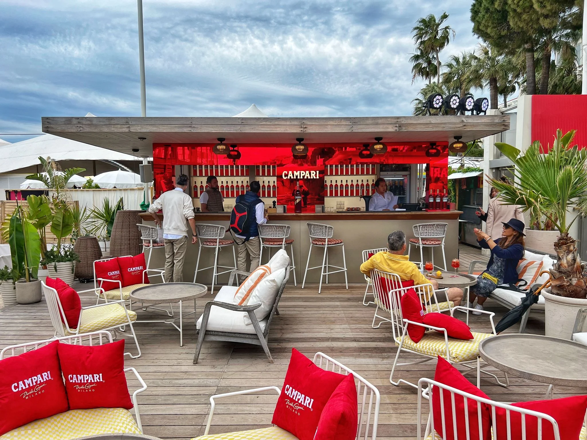 Open-air beach bar with a red Campari backdrop, bar stools and white chairs with red cushions – Campari Cannes Film Festival 2024, France – event architecture