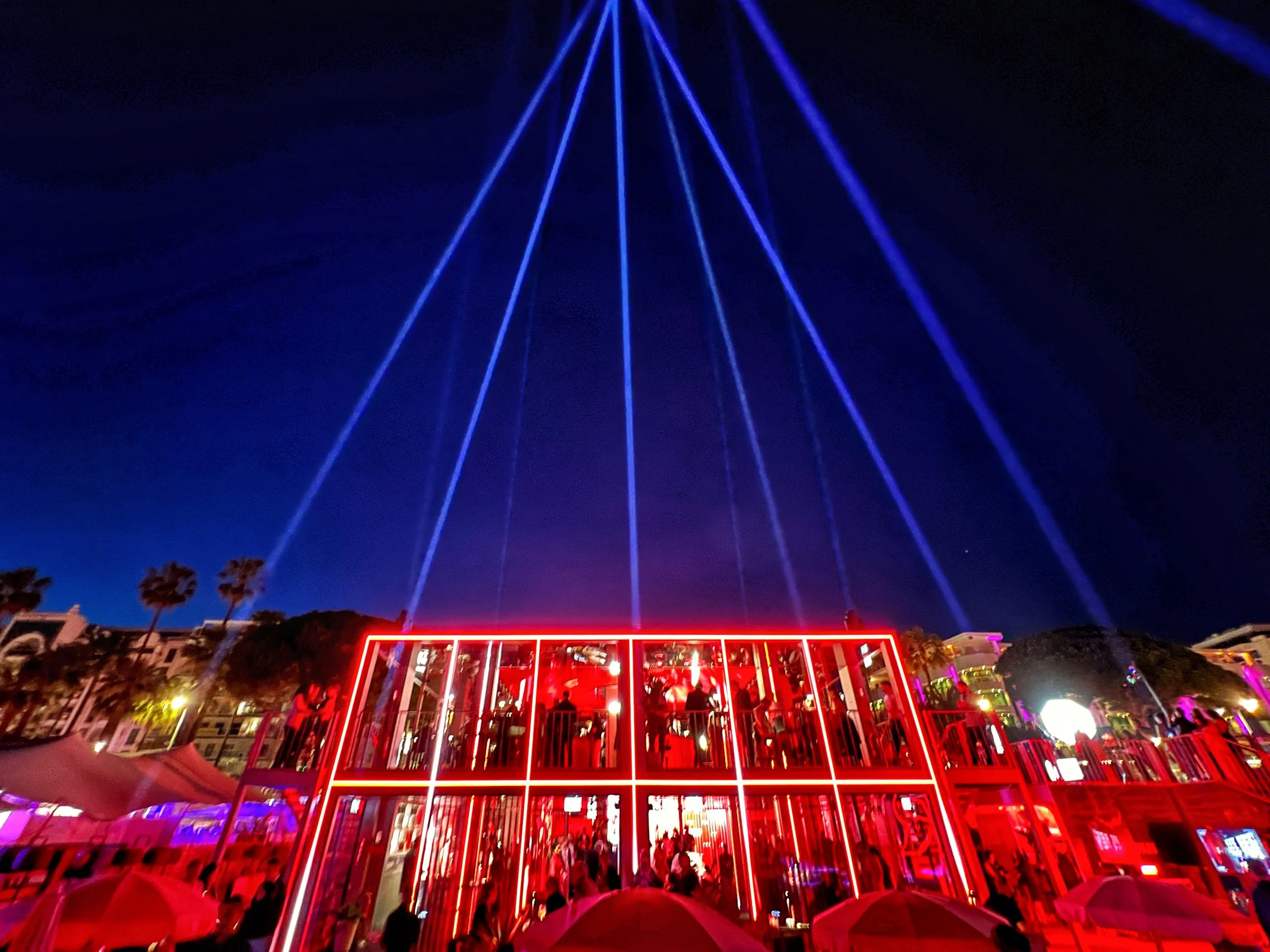 Night view of the pavilion outlined in red light, searchlight beams fanning into the sky above it – Campari Cannes Film Festival 2024, France – event architecture