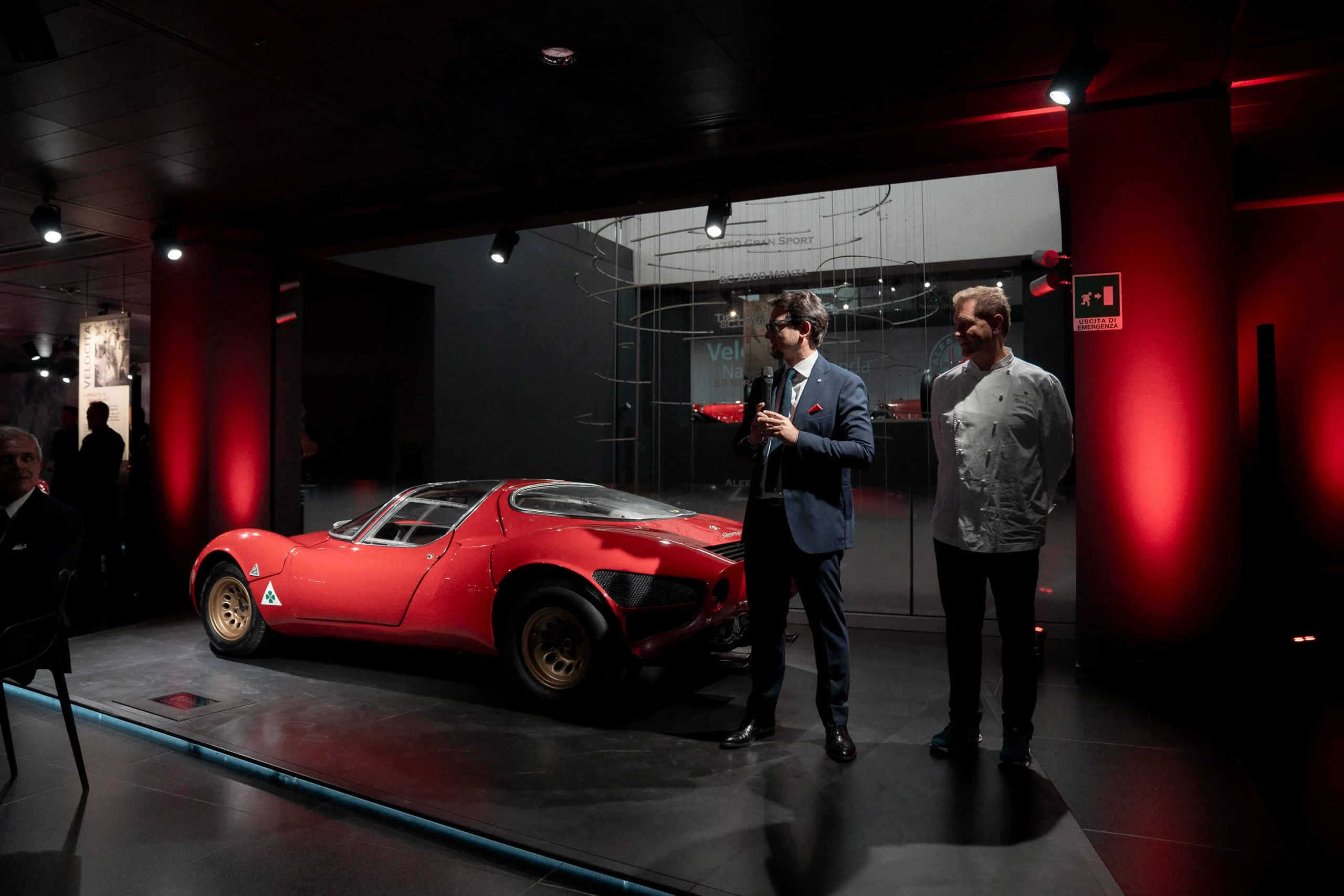 Two speakers next to the original red Alfa Romeo 33 Stradale in a red-lit museum gallery – Alfa Romeo 33 Stradale Unveiling, Arese, Italy, 2023 – event design