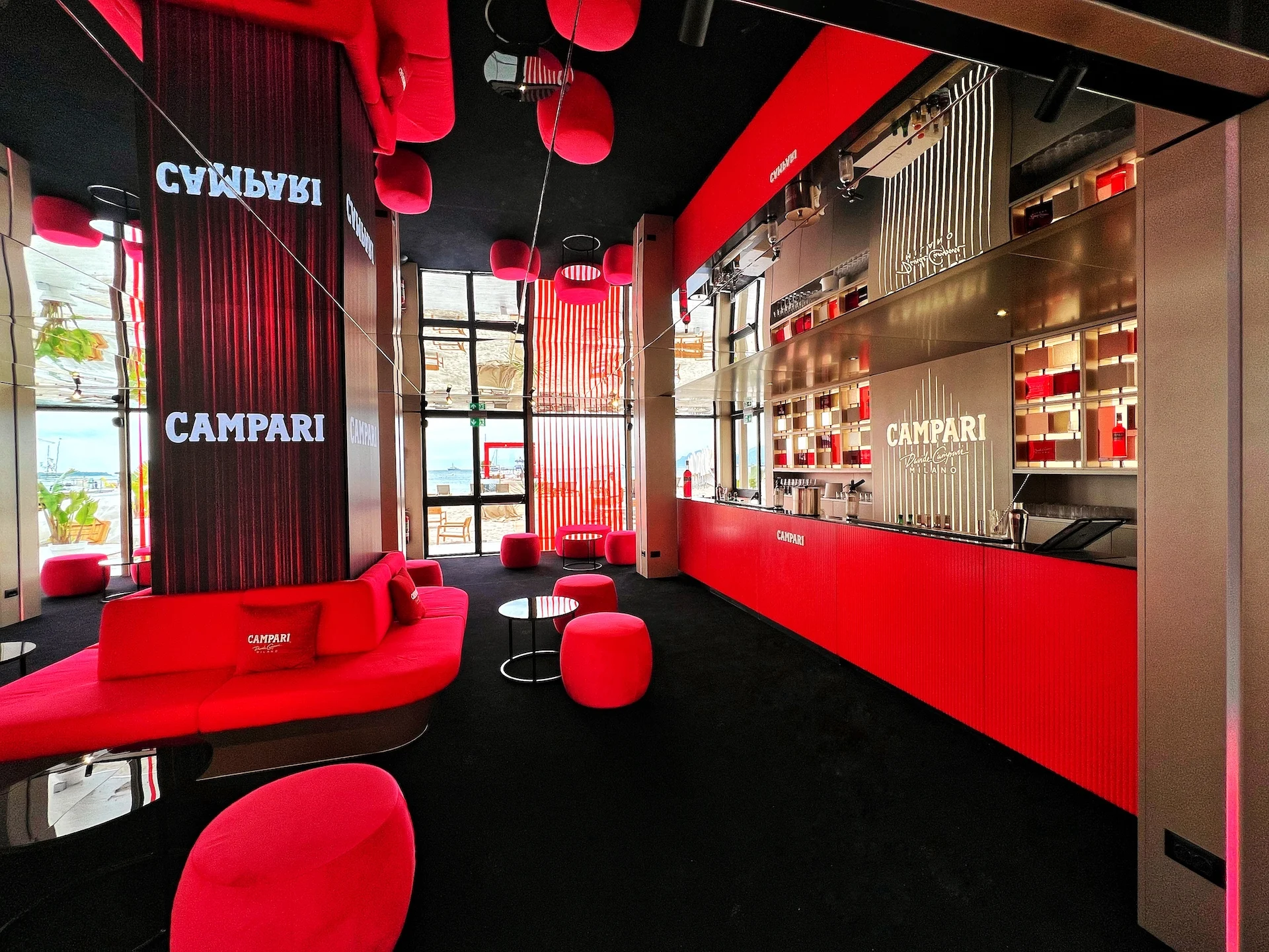 Ground-floor lounge with red sofas and poufs, a Campari-branded column and a long red bar counter – Campari Cannes Film Festival 2024, France – event architecture