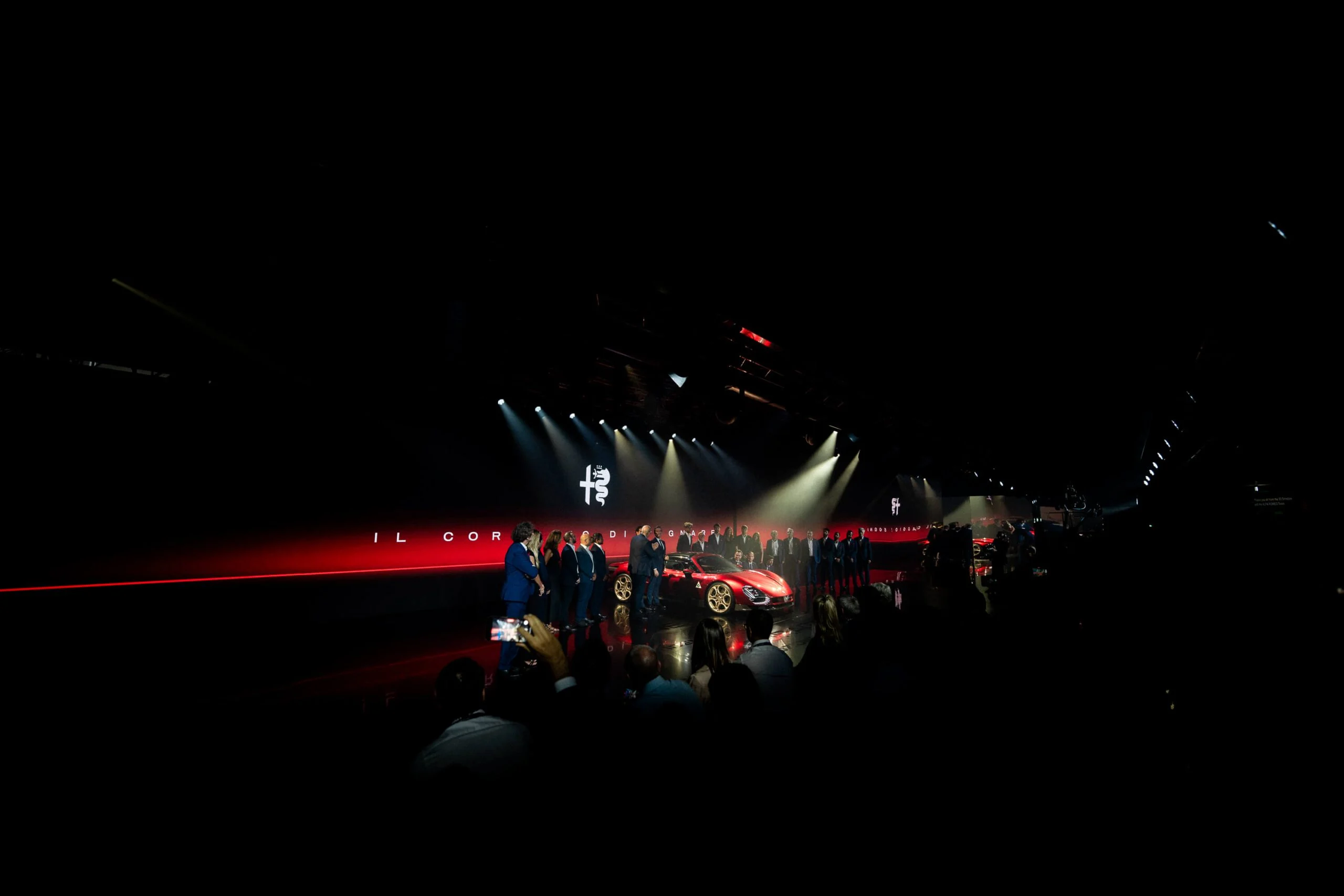 Wide view of the dark hall: guests filming the car on stage beneath the Alfa Romeo logo – Alfa Romeo 33 Stradale Unveiling, Arese, Italy, 2023 – event design