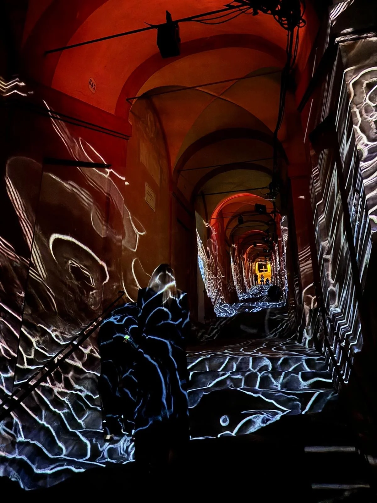 White wave-like projections over the portico walls and steps under red-lit vaults, a visitor in the light – Cesare Cremonini Festival dei Portici, Bologna, Italy, 2024 – event design