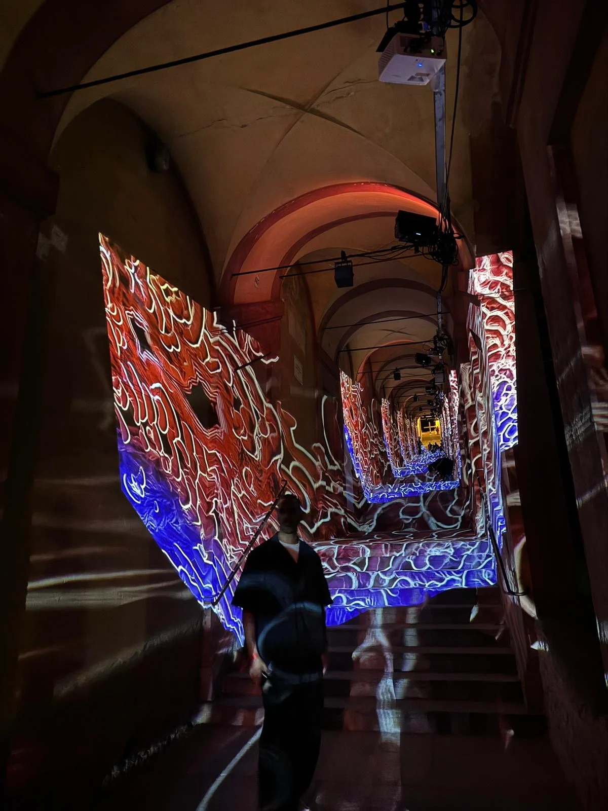 Red and blue organic projections wrapping the portico walls, projectors hanging from the vault, visitor below – Cesare Cremonini Festival dei Portici, Bologna, Italy, 2024 – event design