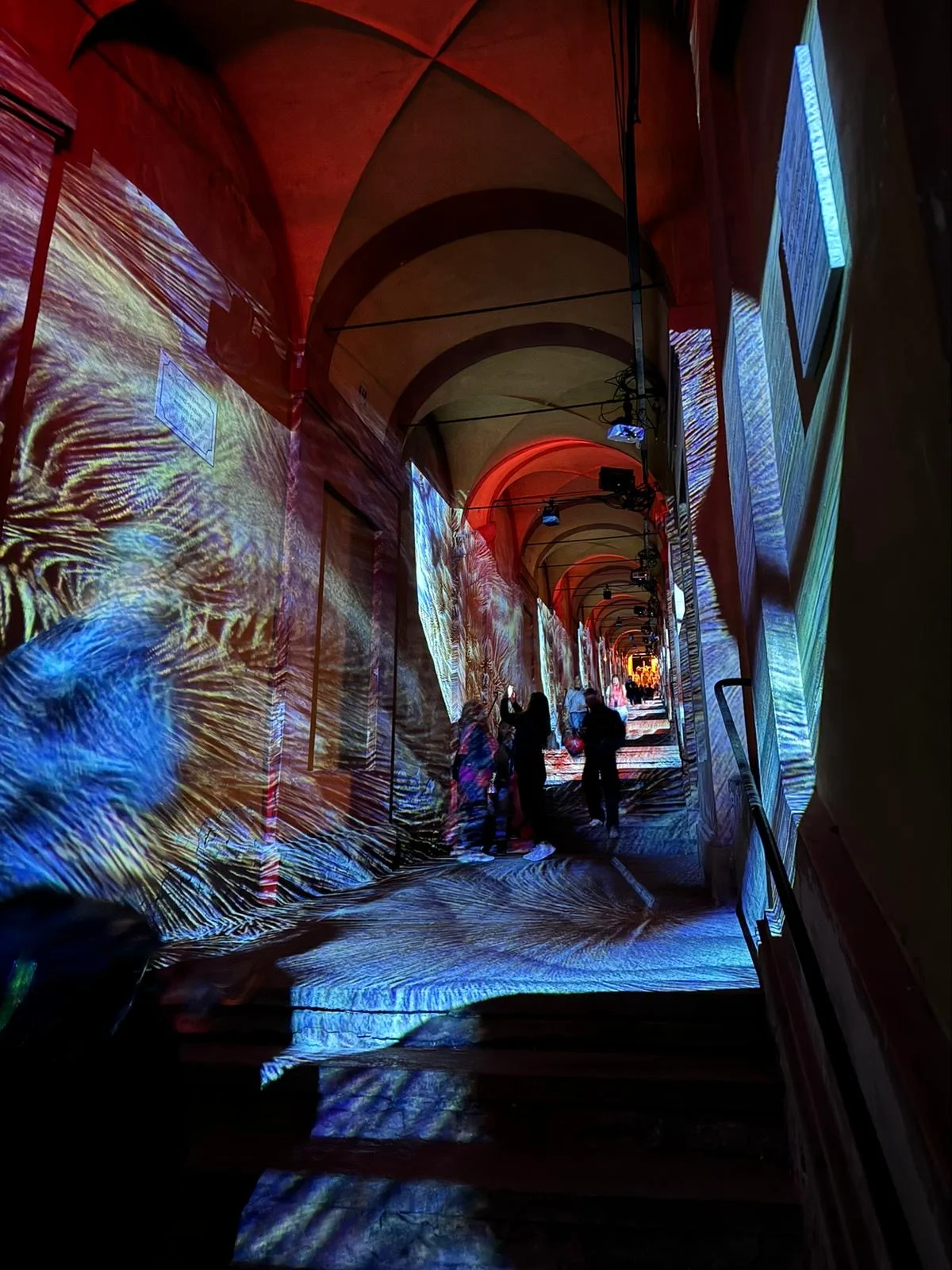 Vaulted San Luca portico with blue and gold swirling projections on walls and steps, visitors walking through – Cesare Cremonini Festival dei Portici, Bologna, Italy, 2024 – event design