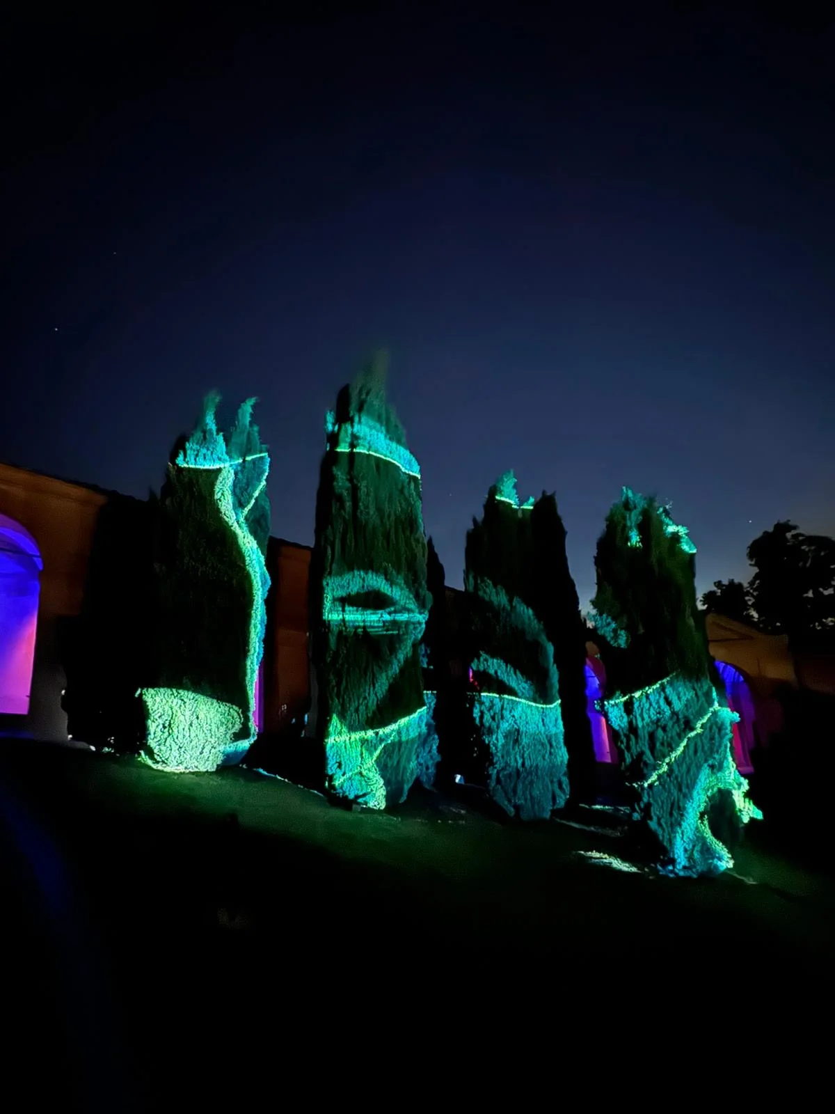 Cypress trees mapped with turquoise light patterns at night, purple-lit arches behind – Cesare Cremonini Festival dei Portici, Bologna, Italy, 2024 – event design