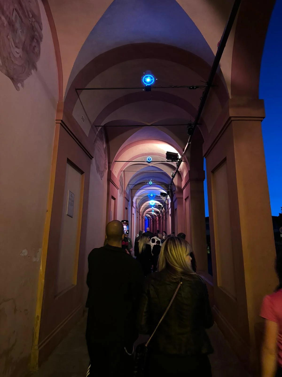 Visitors walking under the portico at dusk, pink-lit vaults with small blue lights along the ceiling – Cesare Cremonini Festival dei Portici, Bologna, Italy, 2024 – event design