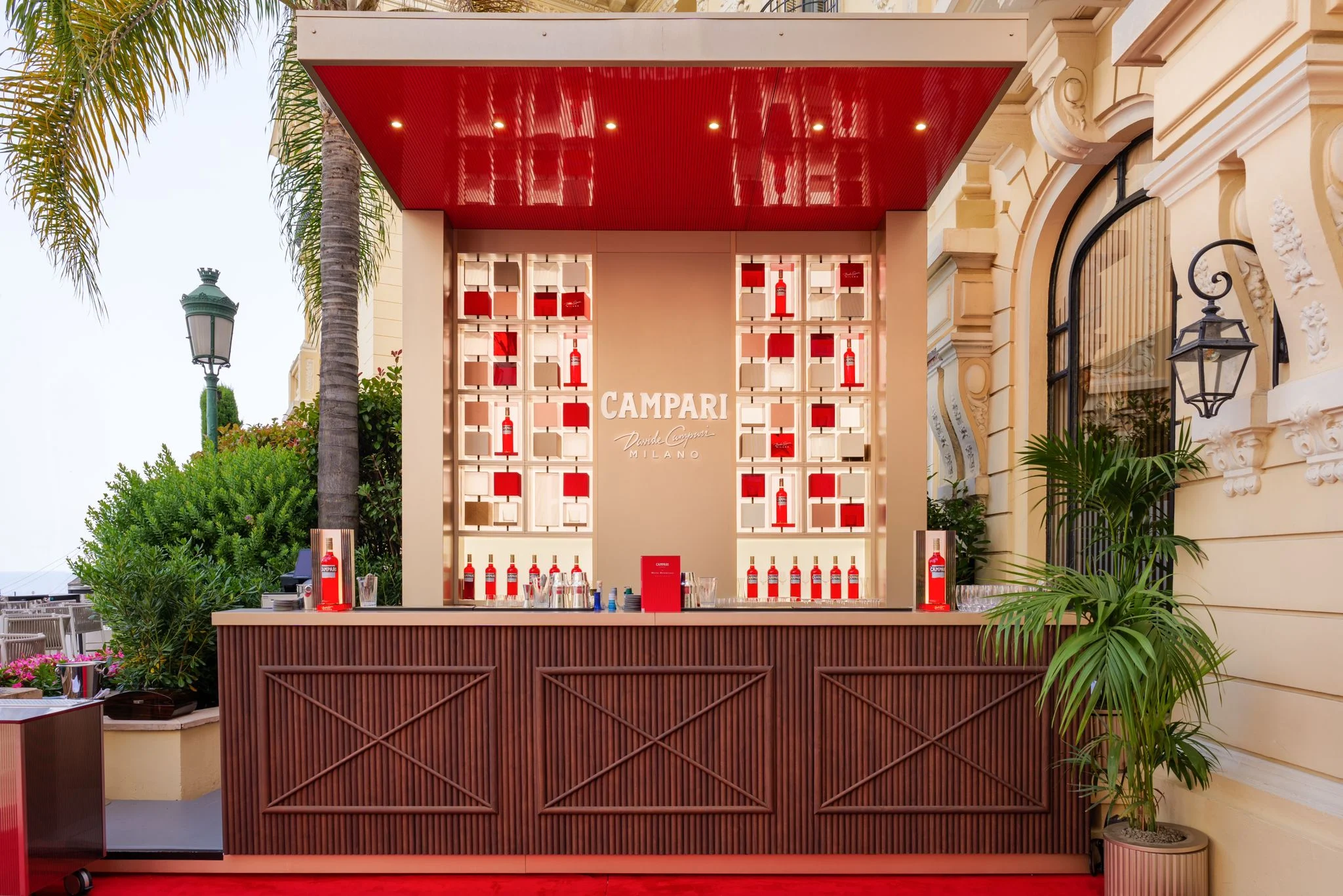 Frontal view of the Campari bar: wooden counter, lit shelves of red bottles and a red lacquered canopy – Campari Pop-Up Bar at the Hôtel Hermitage, Monte Carlo, Monaco, 2024 – hospitality design