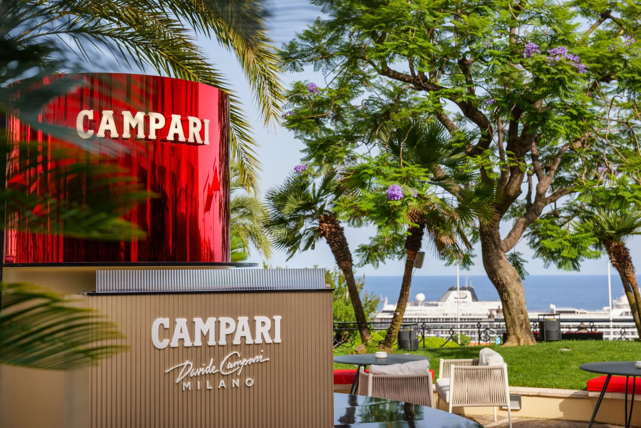 Red mirrored Campari drum above the bar, with garden seating, trees and the sea and harbour behind – Campari Pop-Up Bar at the Hôtel Hermitage, Monte Carlo, Monaco, 2024 – hospitality design