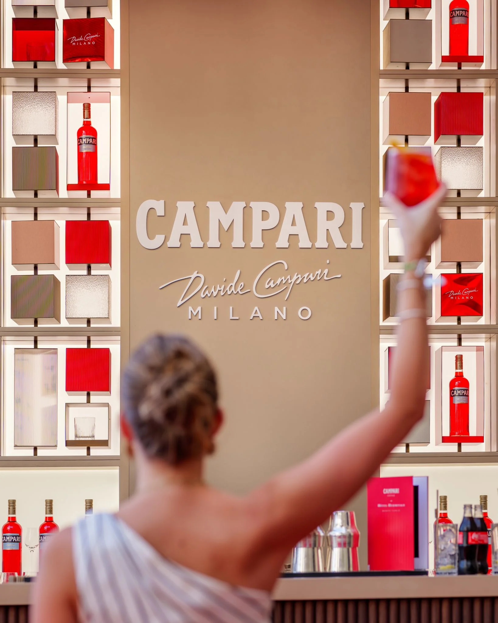 Guest raising a red cocktail in front of the Campari Milano logo wall and shelves of bottles – Campari Pop-Up Bar at the Hôtel Hermitage, Monte Carlo, Monaco, 2024 – hospitality design
