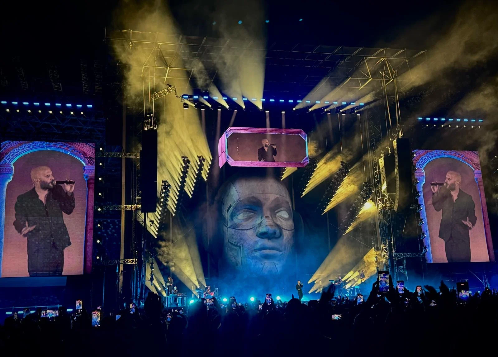 Negramaro stadium stage: giant sculpted head with a visor screen, side LED screens and yellow light beams – Negramaro Stadi 2024, Italy – concert stage design