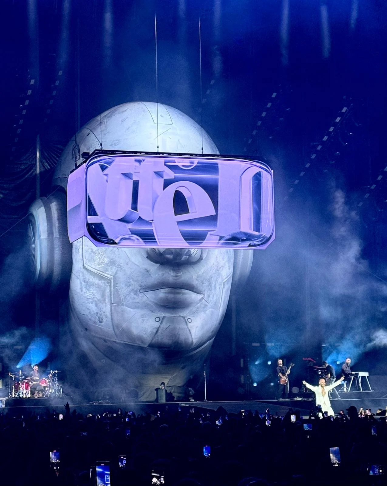 Giant head in blue light, its LED visor showing graphic lettering above the band and singer – Negramaro Stadi 2024, Italy – concert stage design