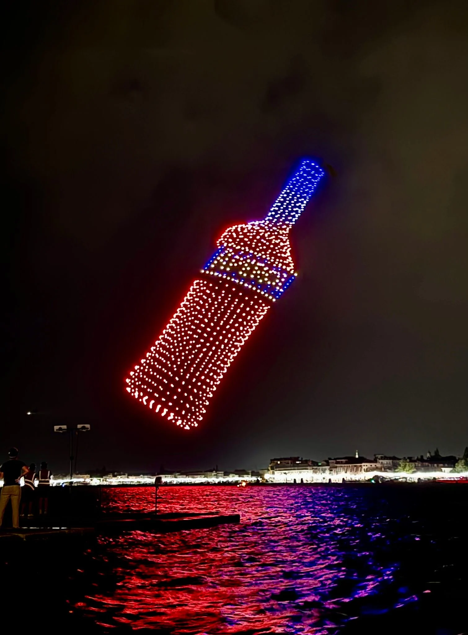 Tilted Aperol bottle drawn by red and blue drones, seen from the waterfront with the lagoon glowing below – Aperol Redentore Drone Show, Venice, Italy, 2024 – creative direction