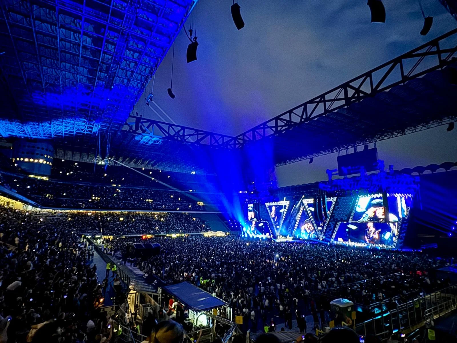 View from the upper stands: blue-lit stadium, crowd on the pitch and the stage screens far away – Vasco Rossi Live 2024, Italy – concert stage design