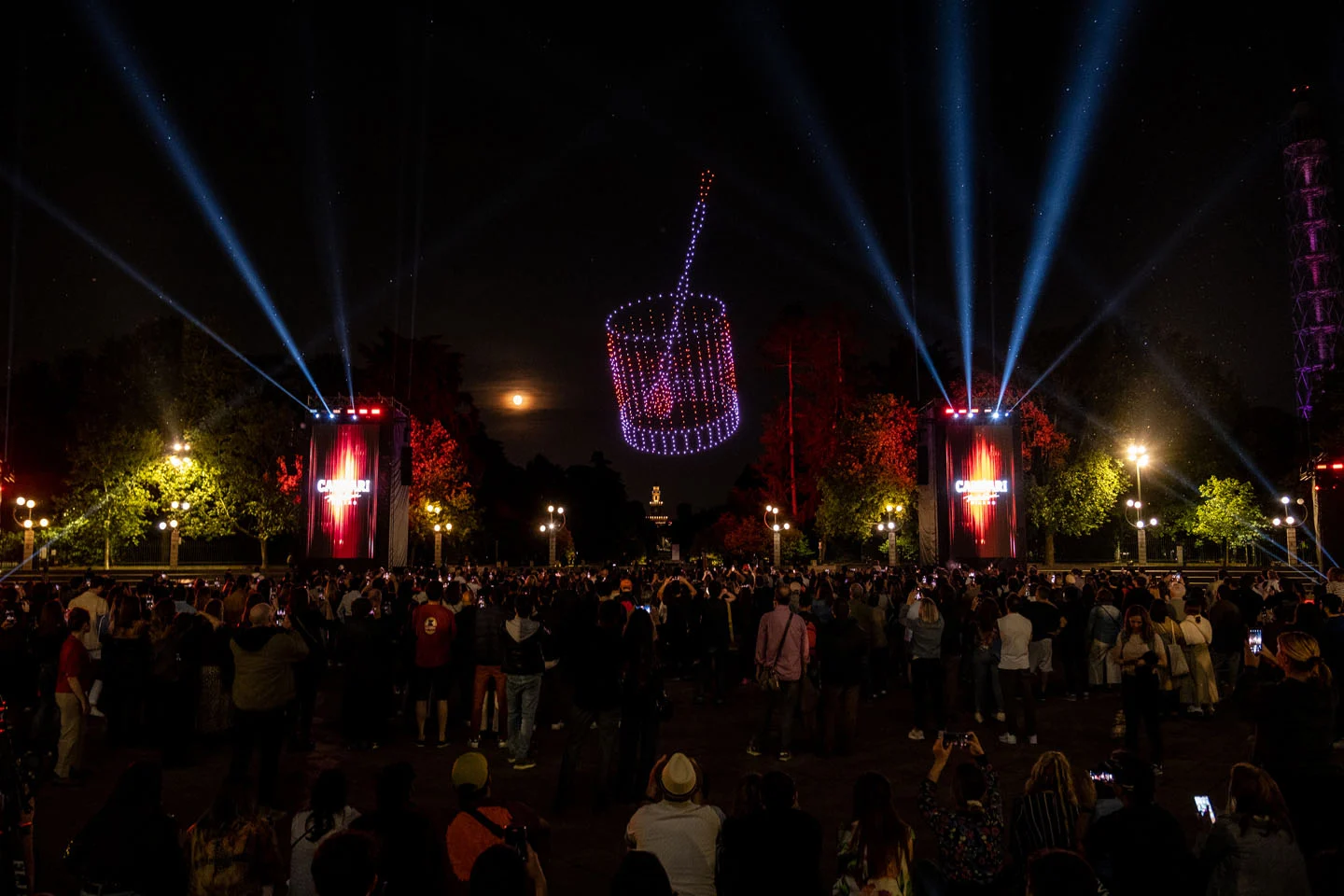 Drones forming a glowing basket shape over a night crowd, searchlights and red Campari towers on either side – Milano House of Campari, Italy, 2023 – event design