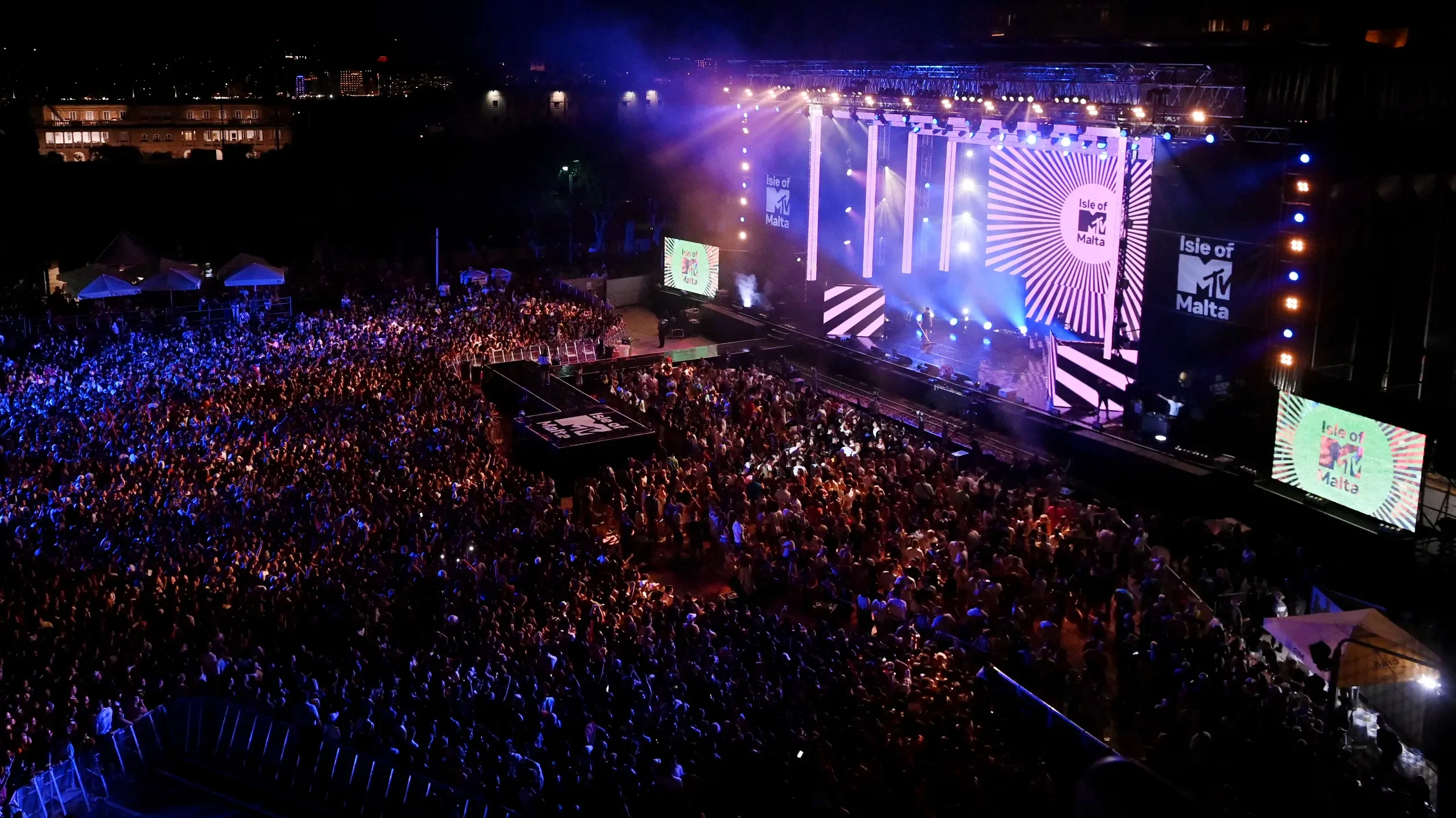Aerial view of the packed Floriana square facing the Isle of MTV stage, side screens and white light rays – Isle of MTV 2024, Floriana, Malta – concert stage design