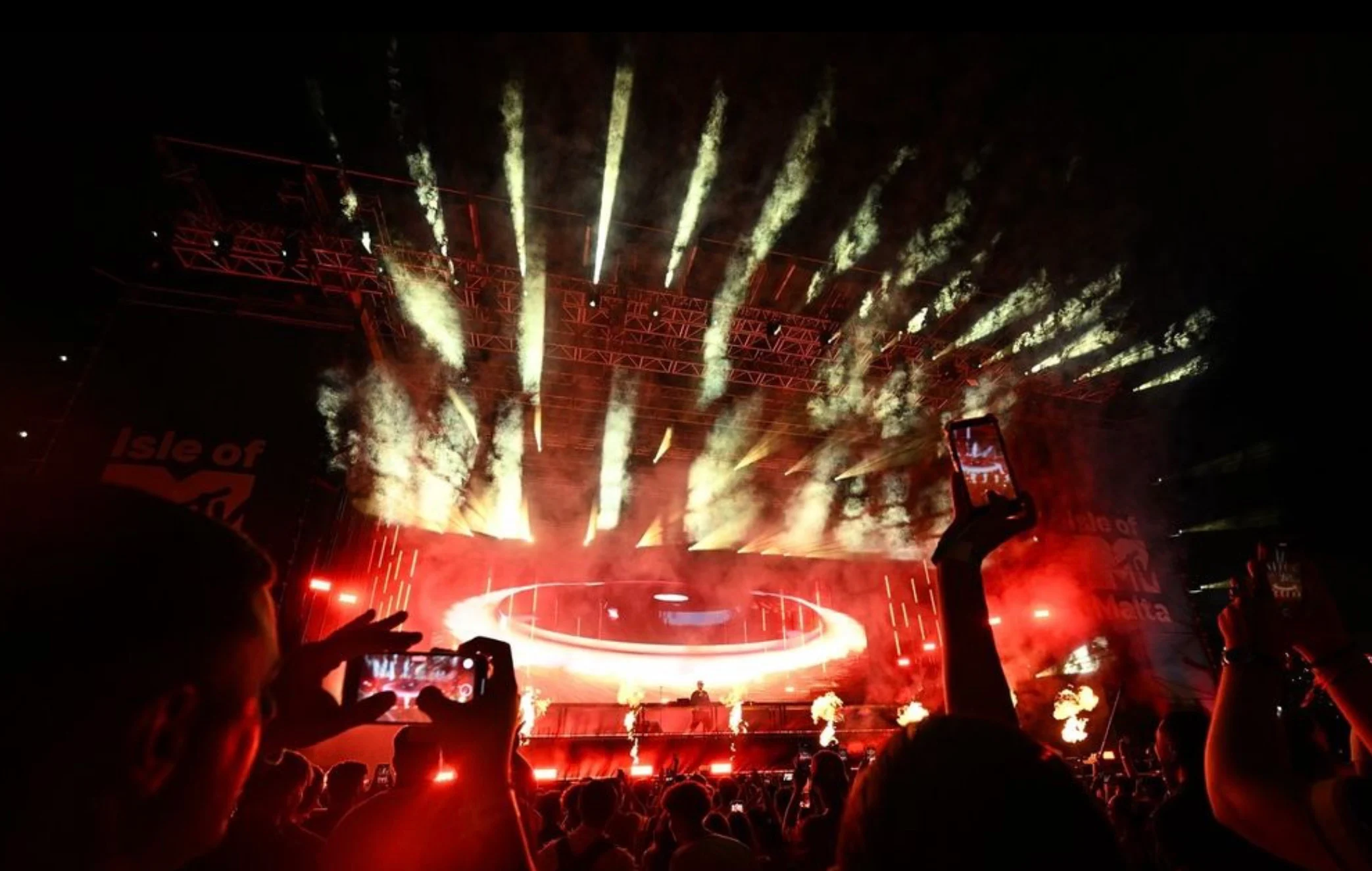 Red-lit stage with a glowing circular ring and fountains of pyrotechnics, fans filming with phones – Isle of MTV 2024, Floriana, Malta – concert stage design