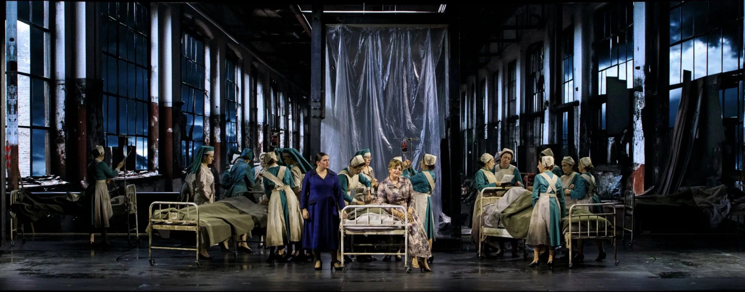 Nurses in pale uniforms around hospital beds inside a derelict industrial hall with tall windows – Il Trovatore at Teatro Comunale Nouveau, Bologna, Italy, 2024 – opera set design