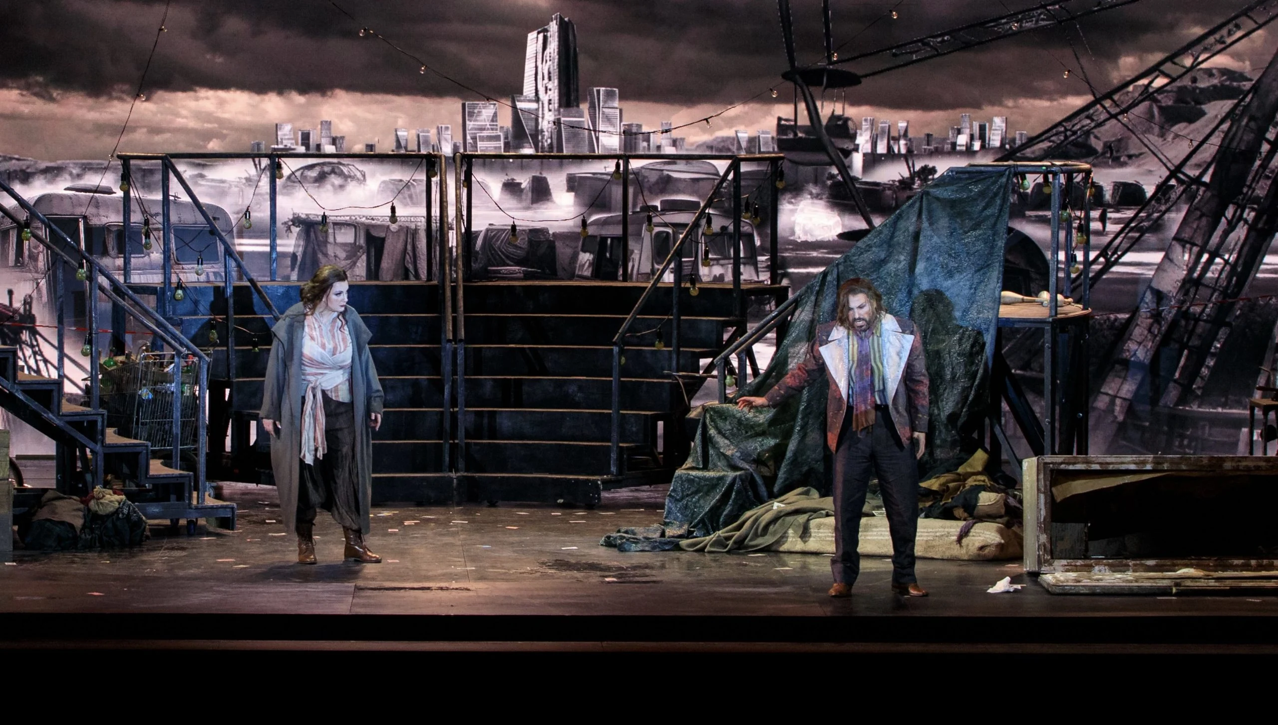 Two singers facing each other on a dark stage with metal stairs, tarpaulins and a ruined city skyline behind – Il Trovatore at Teatro Comunale Nouveau, Bologna, Italy, 2024 – opera set design