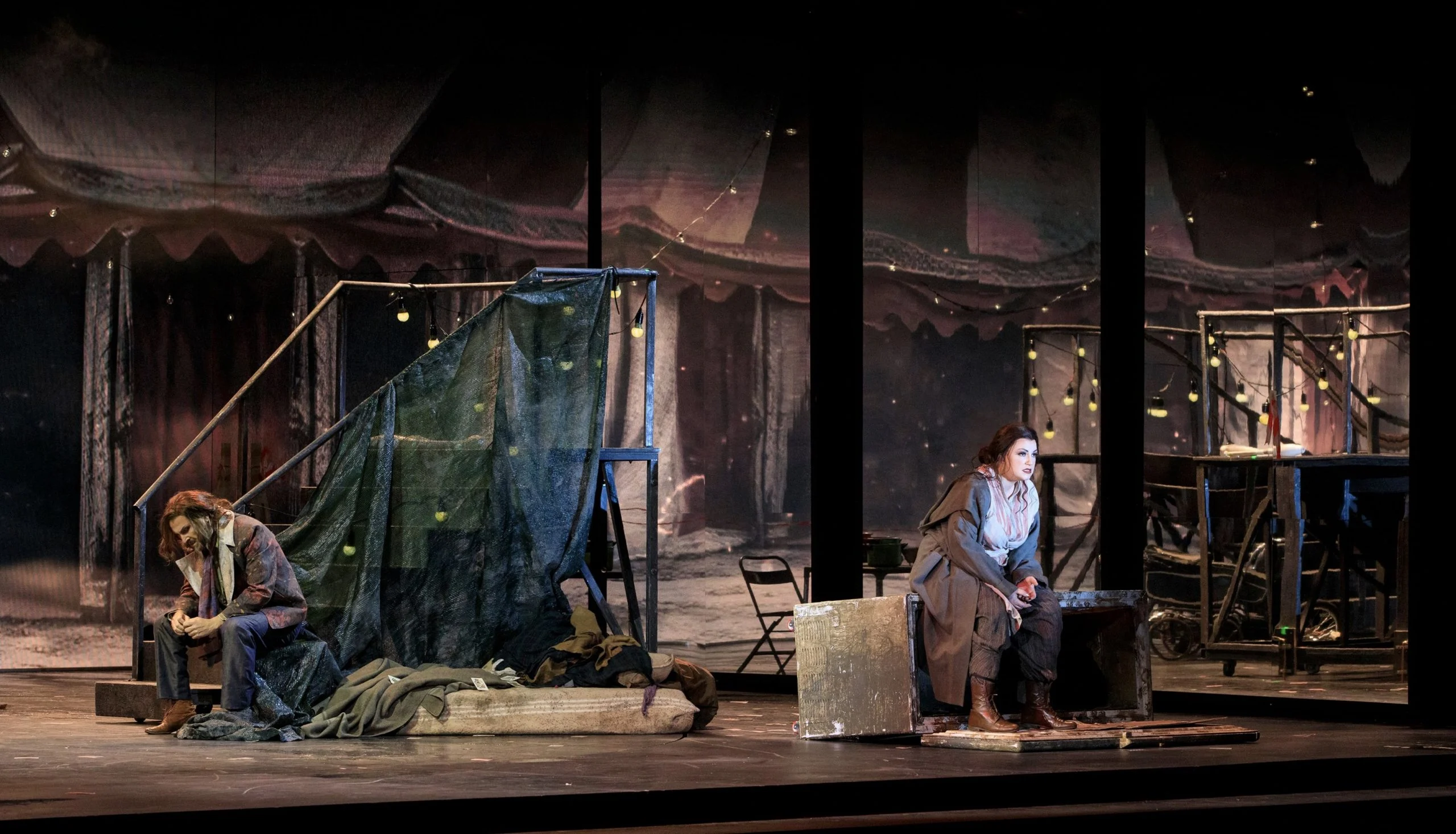 Two singers seated apart at night amid a circus set of tents, fairy lights and metal scaffolding – Il Trovatore at Teatro Comunale Nouveau, Bologna, Italy, 2024 – opera set design