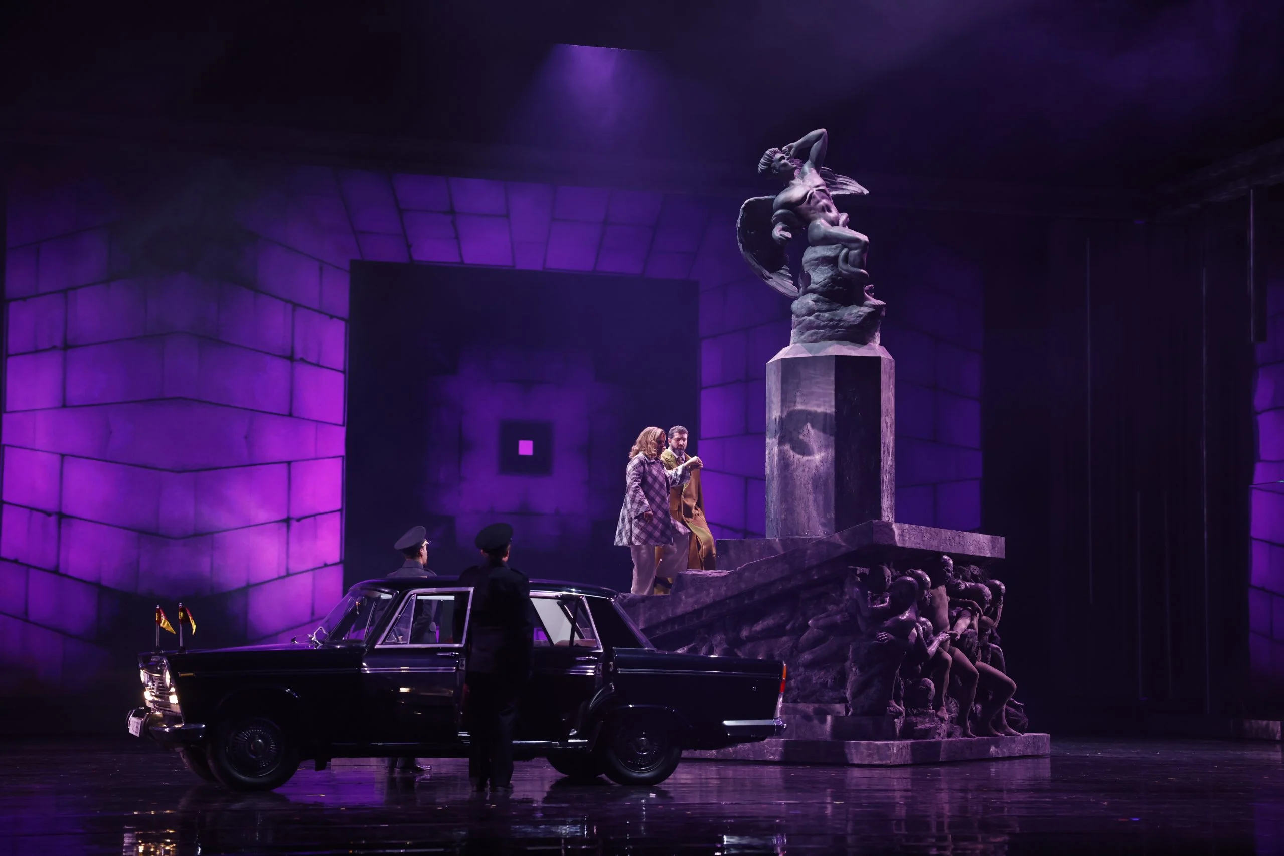 Two singers on the steps of a sculpted monument bathed in purple light, a black vintage car and guards below – Don Carlo at the Royal Danish Theatre, Copenhagen, Denmark, 2024 – opera set design