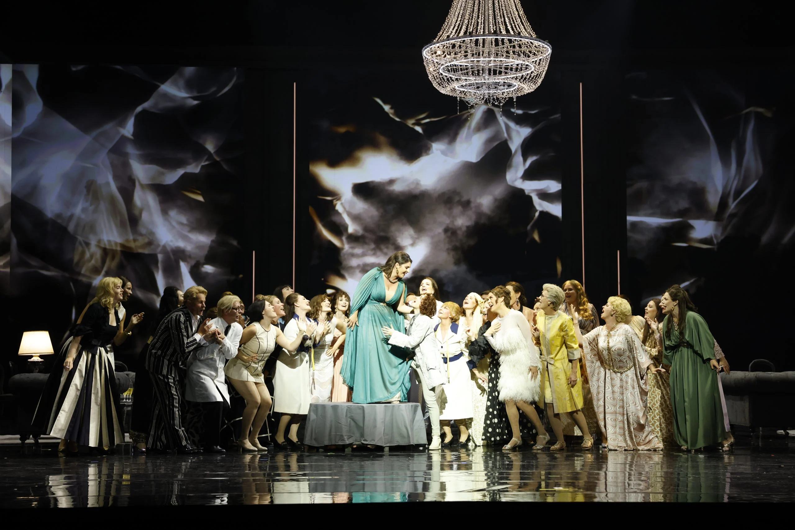 Women's chorus in colourful dresses around a singer in green standing on a table, under a crystal chandelier – Don Carlo at the Royal Danish Theatre, Copenhagen, Denmark, 2024 – opera set design