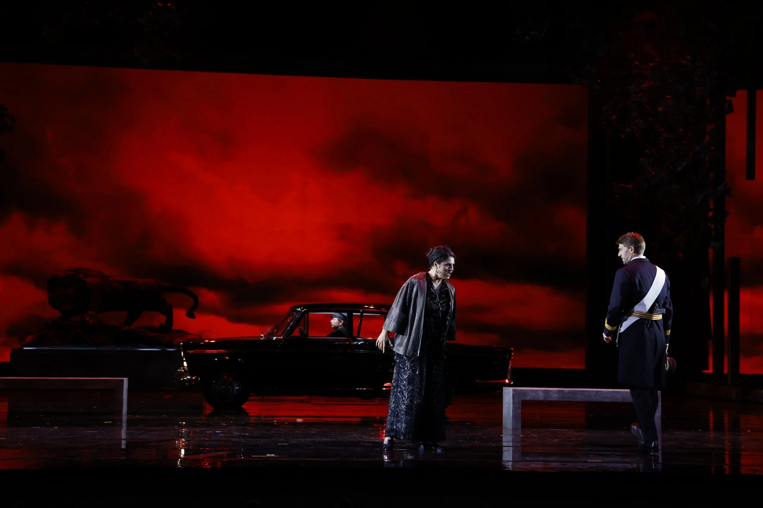 Singer in black before a blazing red sky projection, an officer in uniform and a black car behind – Don Carlo at the Royal Danish Theatre, Copenhagen, Denmark, 2024 – opera set design